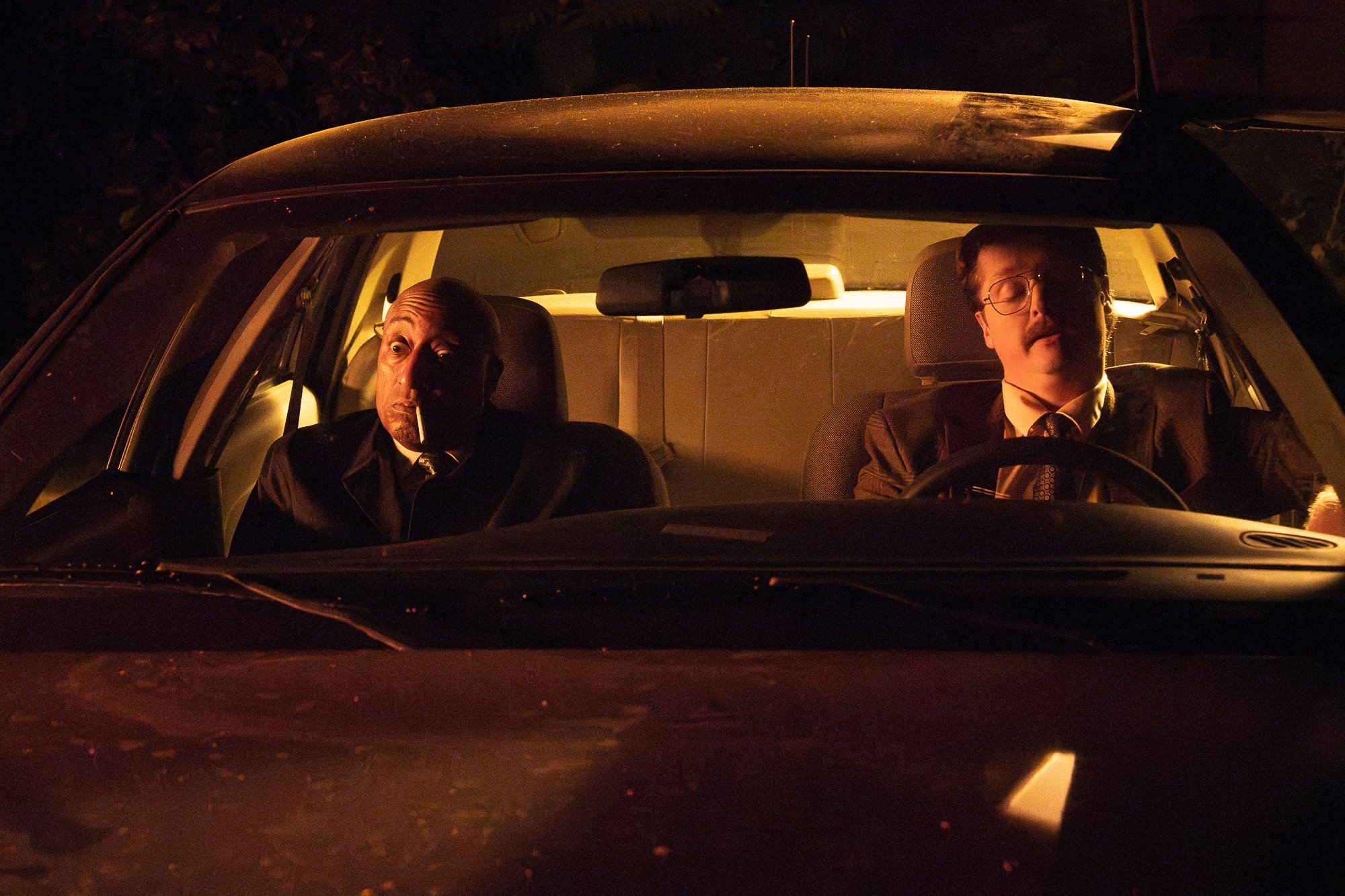 Two men are sitting in a car at night. One man is in the passenger seat, smoking a cigarette, bald with a serious expression, wearing a suit and tie. The other man is in the driver's seat, wearing glasses, also dressed in a suit and tie, with a serio