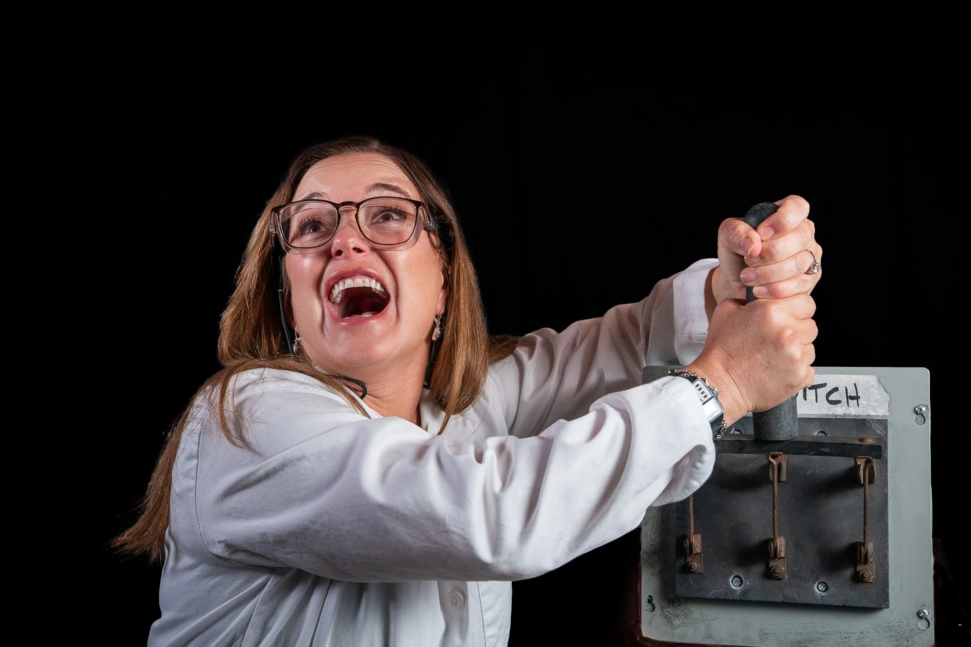 Woman with glasses and a white shirt appears to be pulling a large lever in a black background setting.