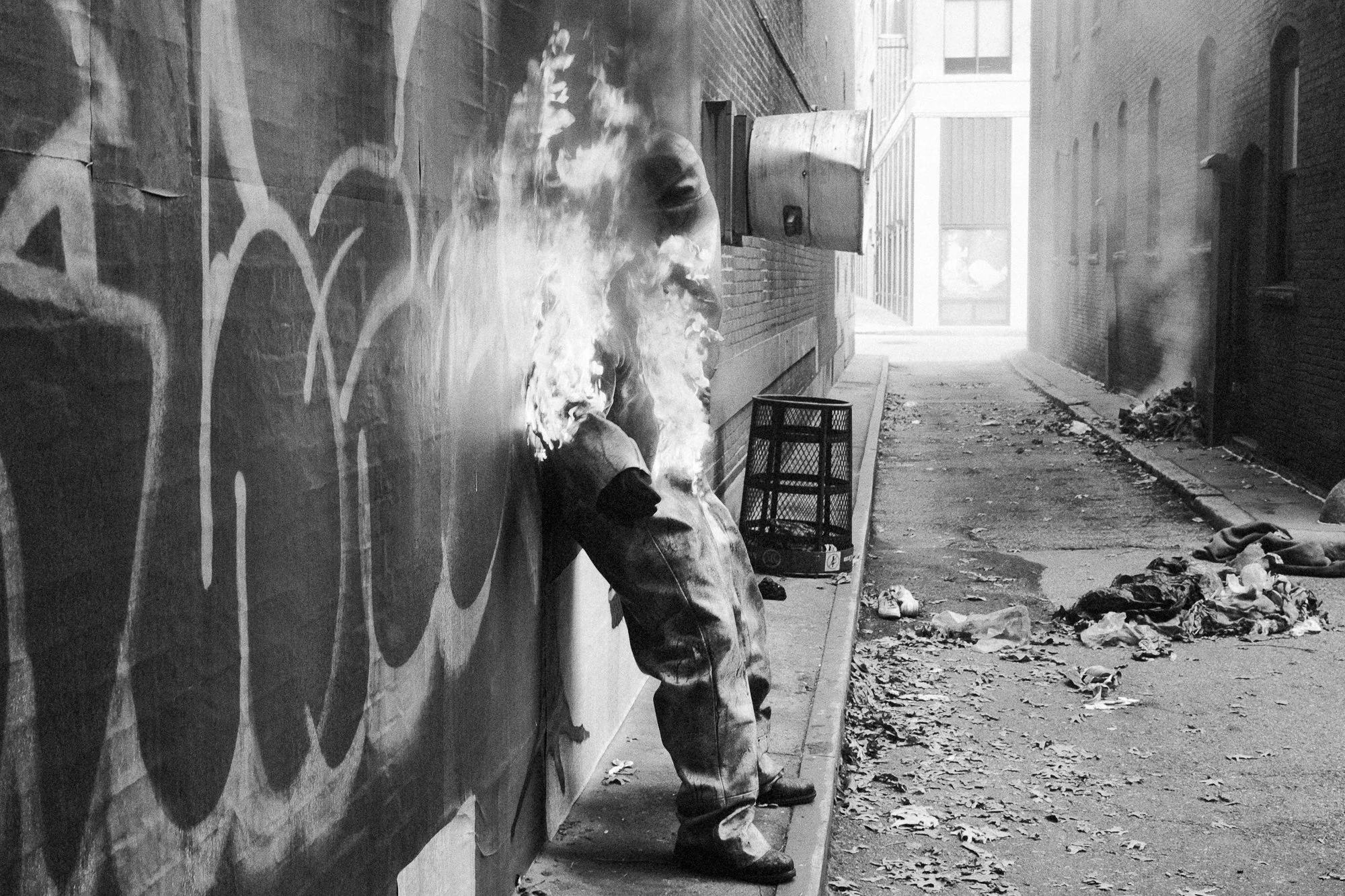 A person sitting on a sidewalk against a graffiti-covered wall, smoking a cigarette, with trash and debris scattered on the ground nearby.