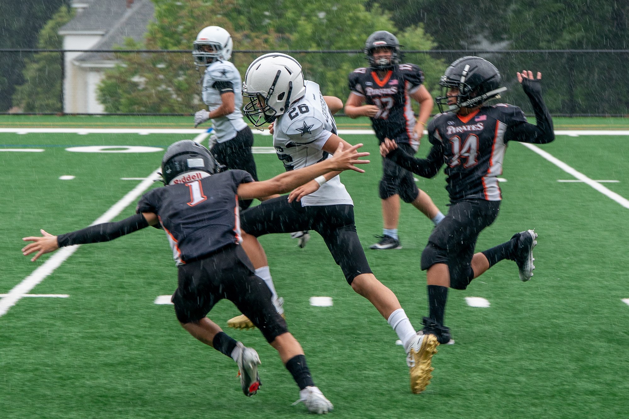 A group of young football players in rain, playing a game on a field. One player in white jersey with the number 26 is holding the football, while others in black jerseys with numbers 14 and 1 are reaching or blocking.