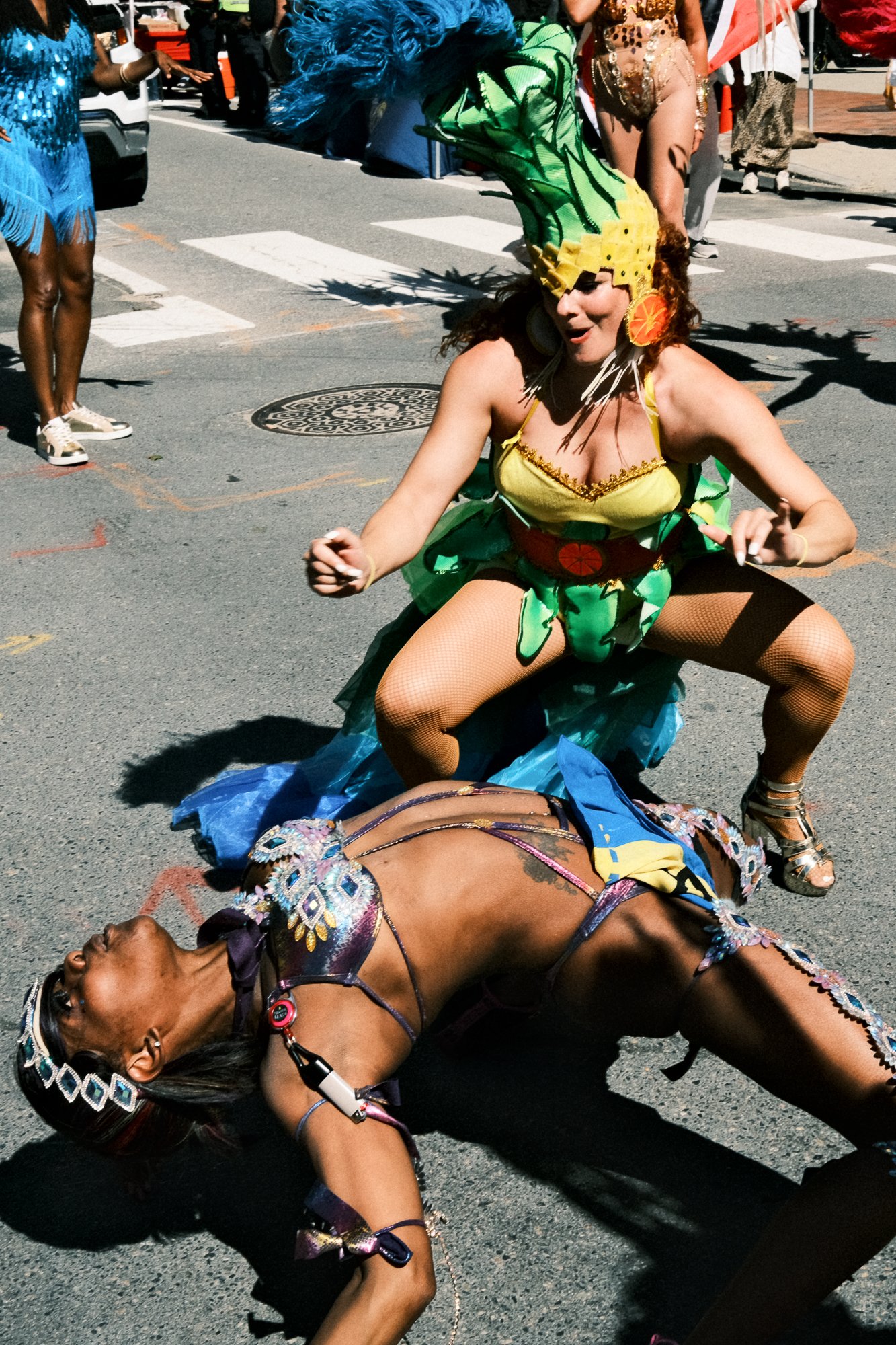 People participating in a parade or festival, dressed in colorful and elaborate costumes, with one woman crouching on the street in a vibrant feathered headdress and costume, others standing around wearing similar attire.