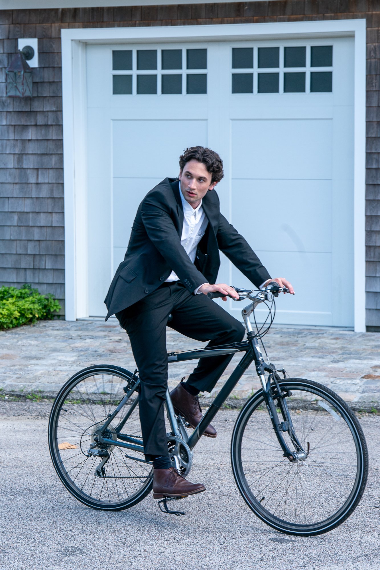A young man in formal attire, including a black suit and brown shoes, is riding a black bicycle in front of a white garage door with a wooden exterior. The scene is outdoors on a paved surface, with some greenery on the side.