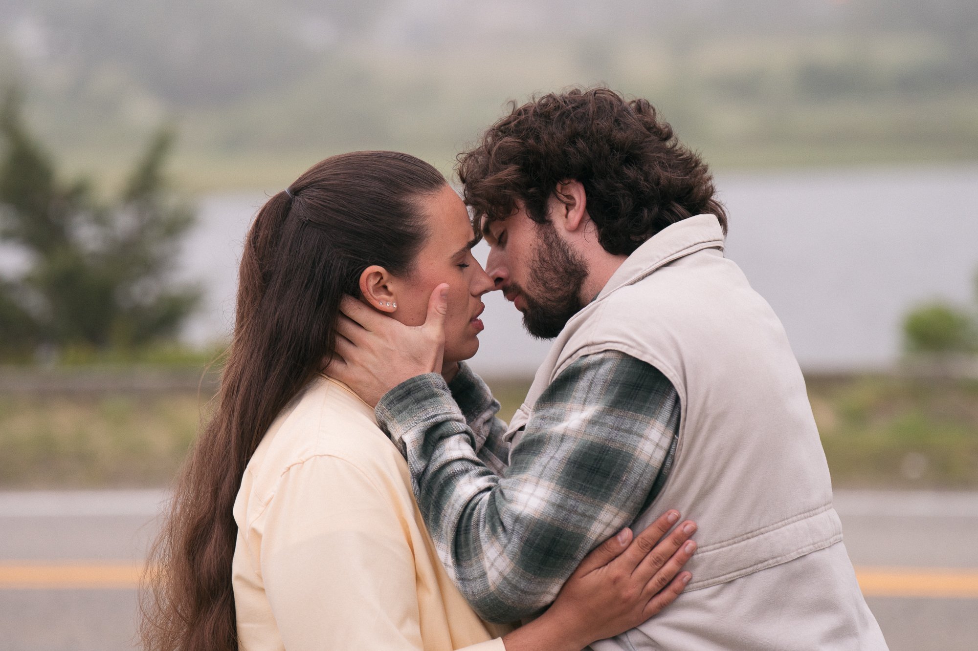A young man and woman are close together outdoors, with their foreheads touching, eyes closed, and holding each other's faces and waists in an intimate moment.