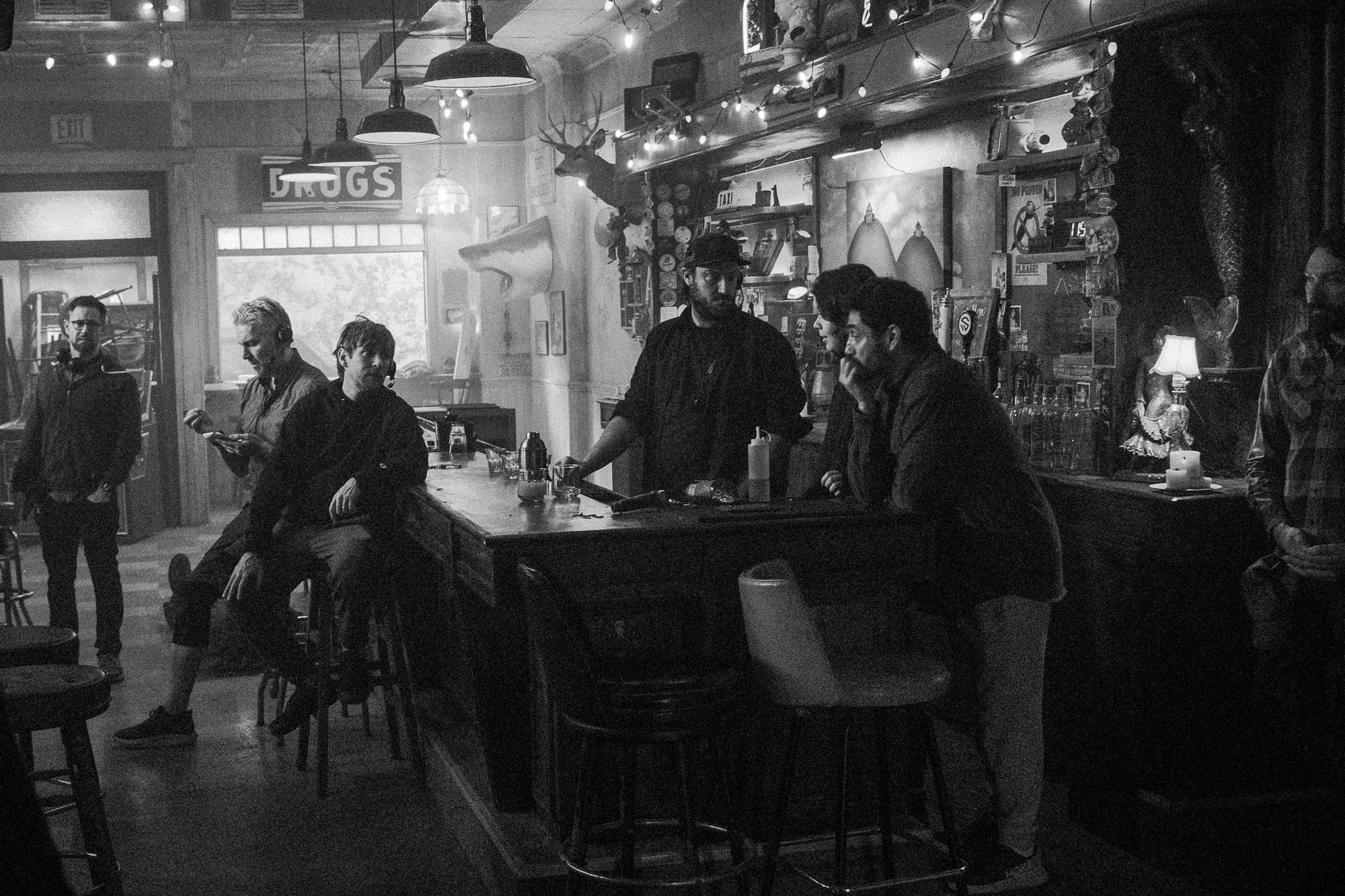 Black and white photo of a bar with several people interacting. Some are standing, others are seated around the bar. The bar has various bottles and decorations, including a mounted deer head on the wall. There are string lights and hanging lamps, cr