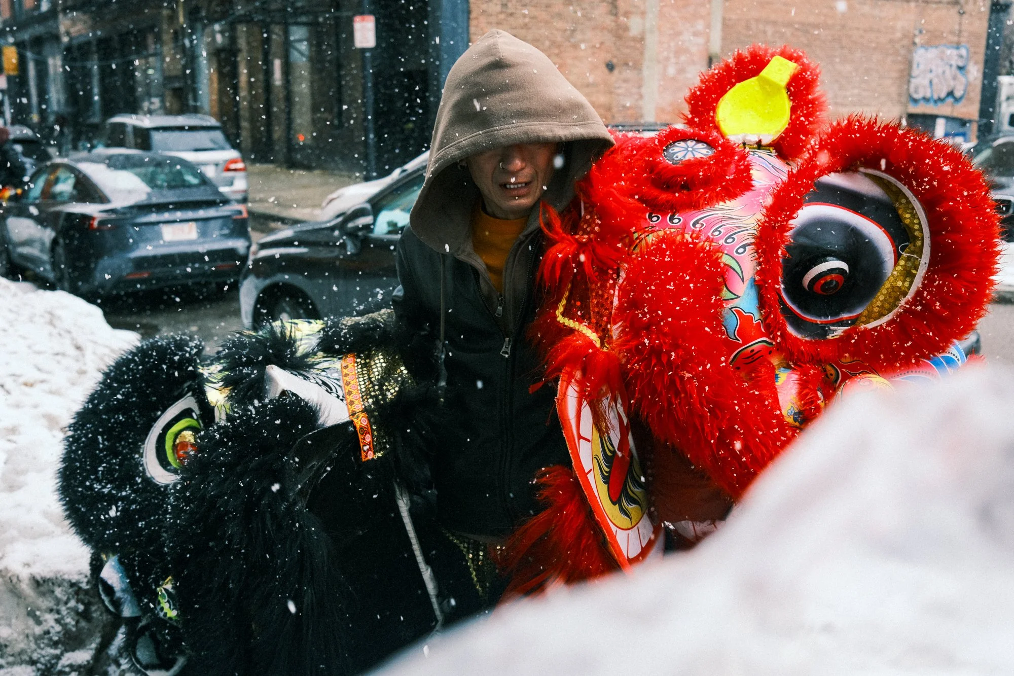 A person in a hooded jacket standing next to a colorful dragon dance costume in a snowy urban street.