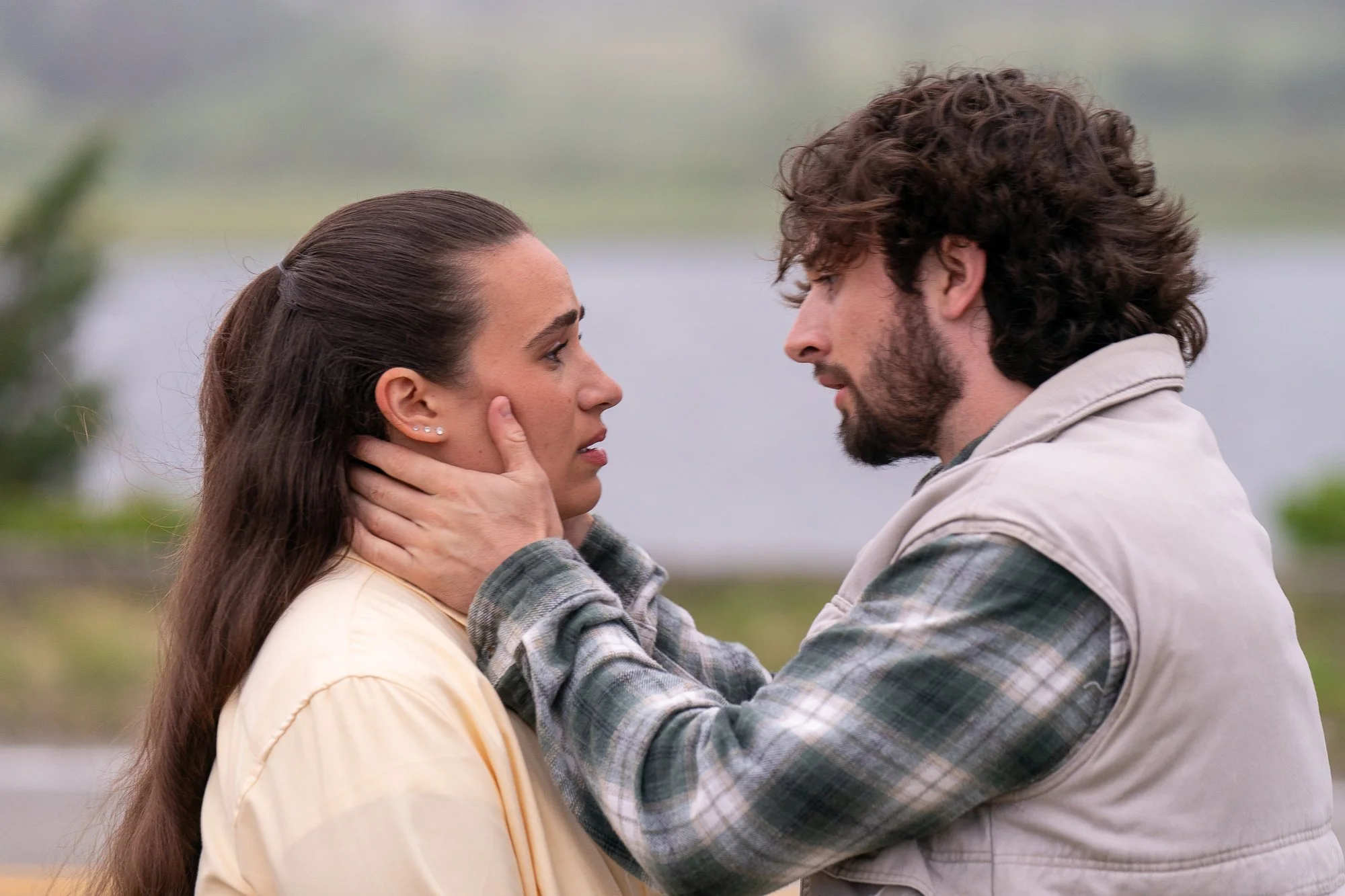 A man and woman are standing close together outdoors, gazing into each other's eyes with the man holding the woman's face gently. The woman has long brown hair and the man has curly hair and a beard, both dressed casually.