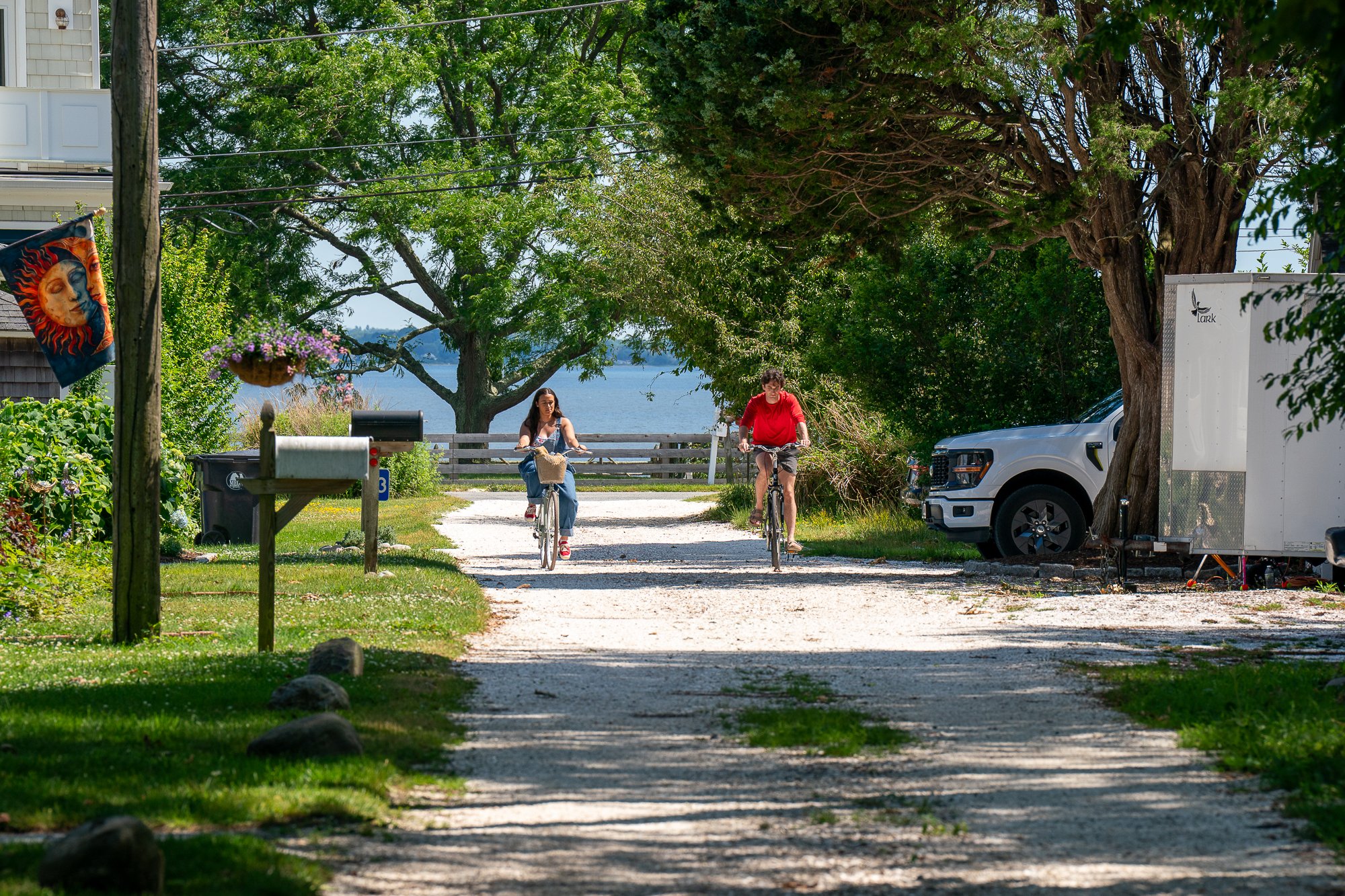 Two women riding bicycles on a gravel path next to trees and a body of water in the background. One woman wears a black shirt and jeans with a basket on her bike; the other wears a red shirt. A white truck is parked to the right, with a large tree be
