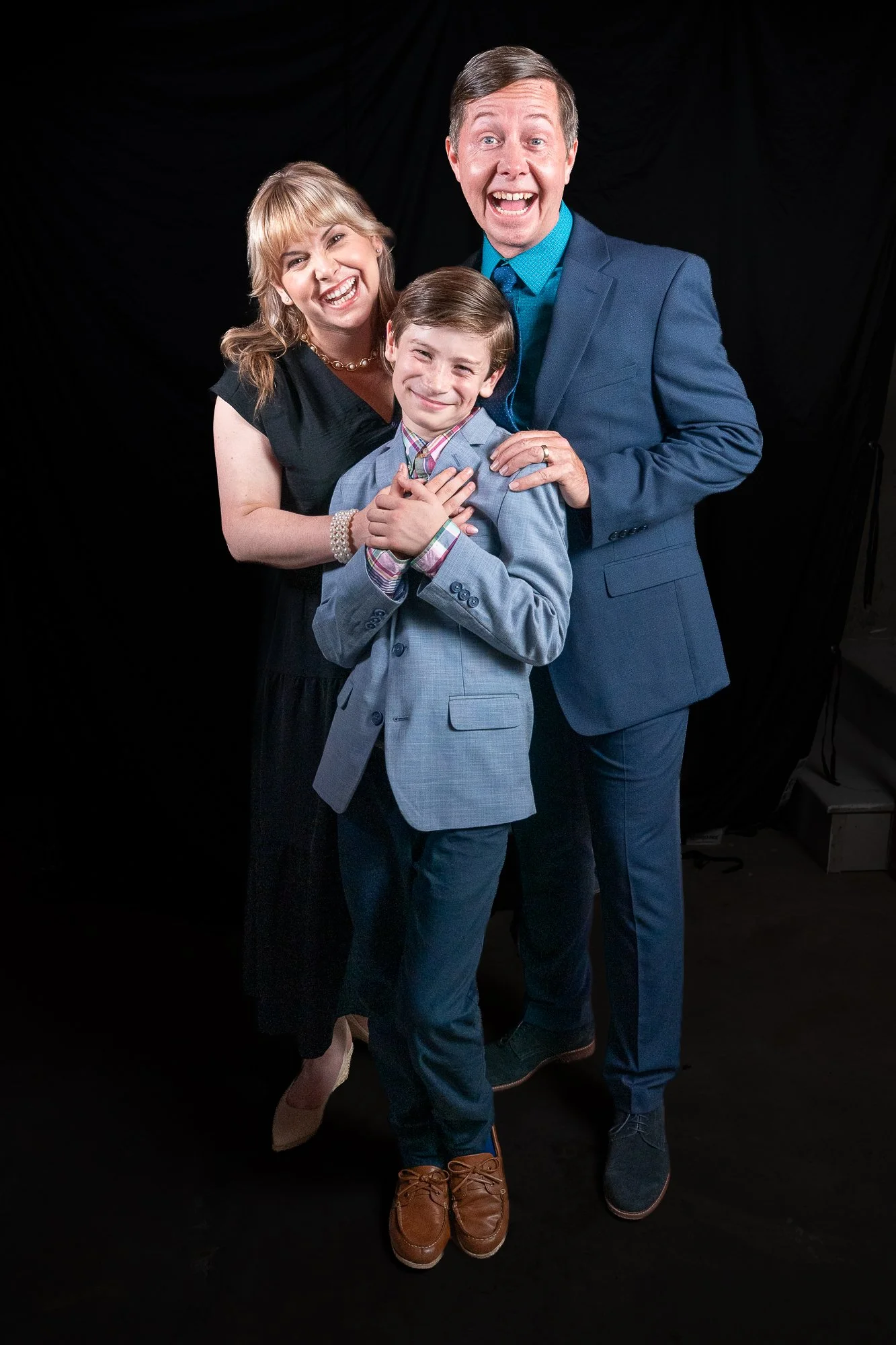 Three people, a man, a woman, and a boy, smiling and standing close together against a black background. The woman is on the left, the boy in the center, and the man on the right.
