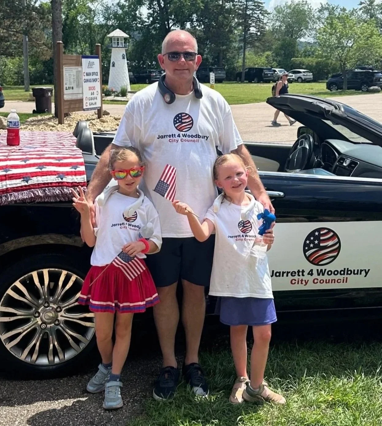 A man in sunglasses and a white campaign T-shirt stands with two young girls, all holding American flags. They are outdoors near a vehicle with a logo for Jarrett 4 Woodbury City Council, surrounded by trees and parked cars.