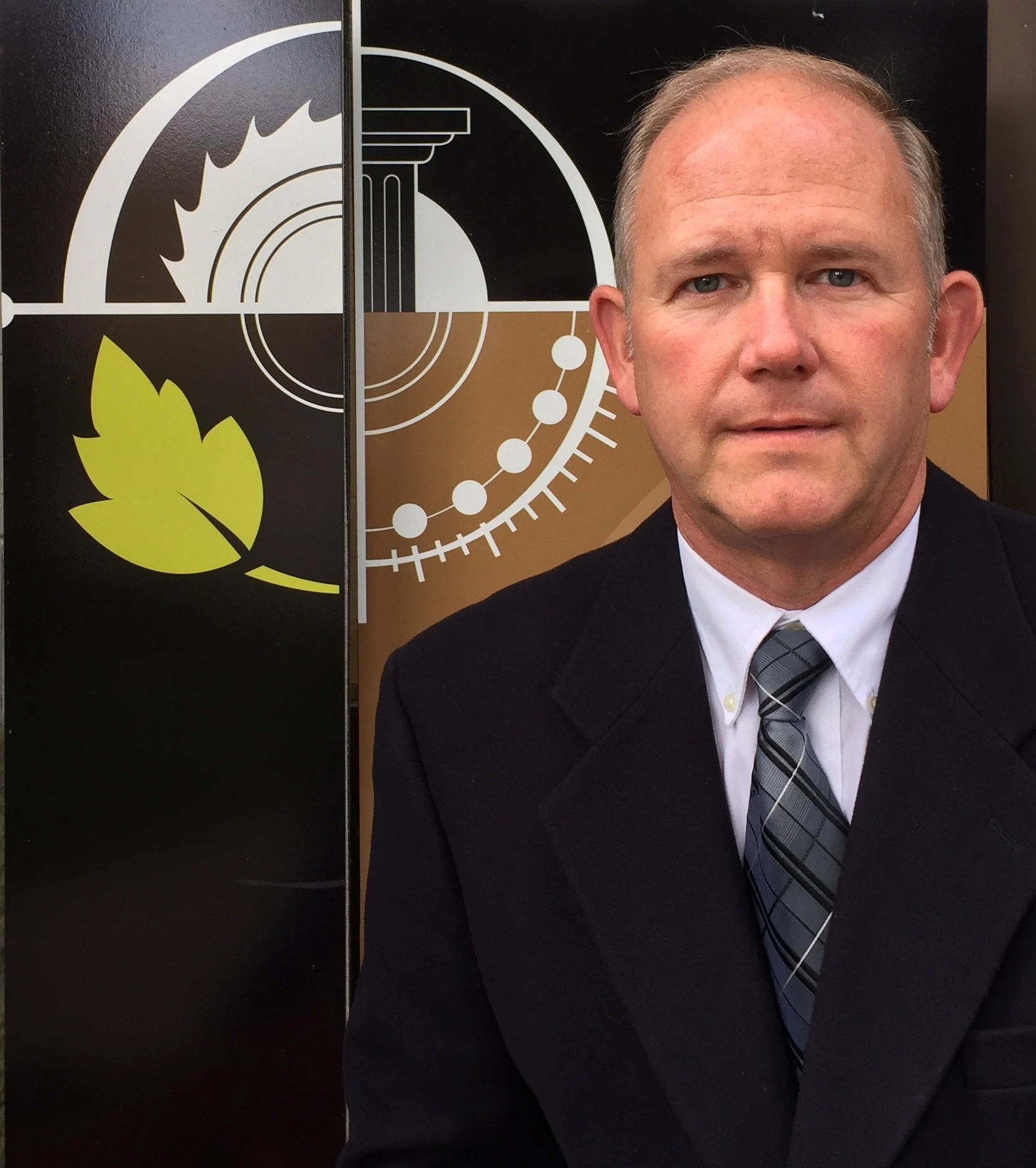 A man in a business suit and a diagonally striped tie standing in front of a black and brown background with circular and linear graphic designs.