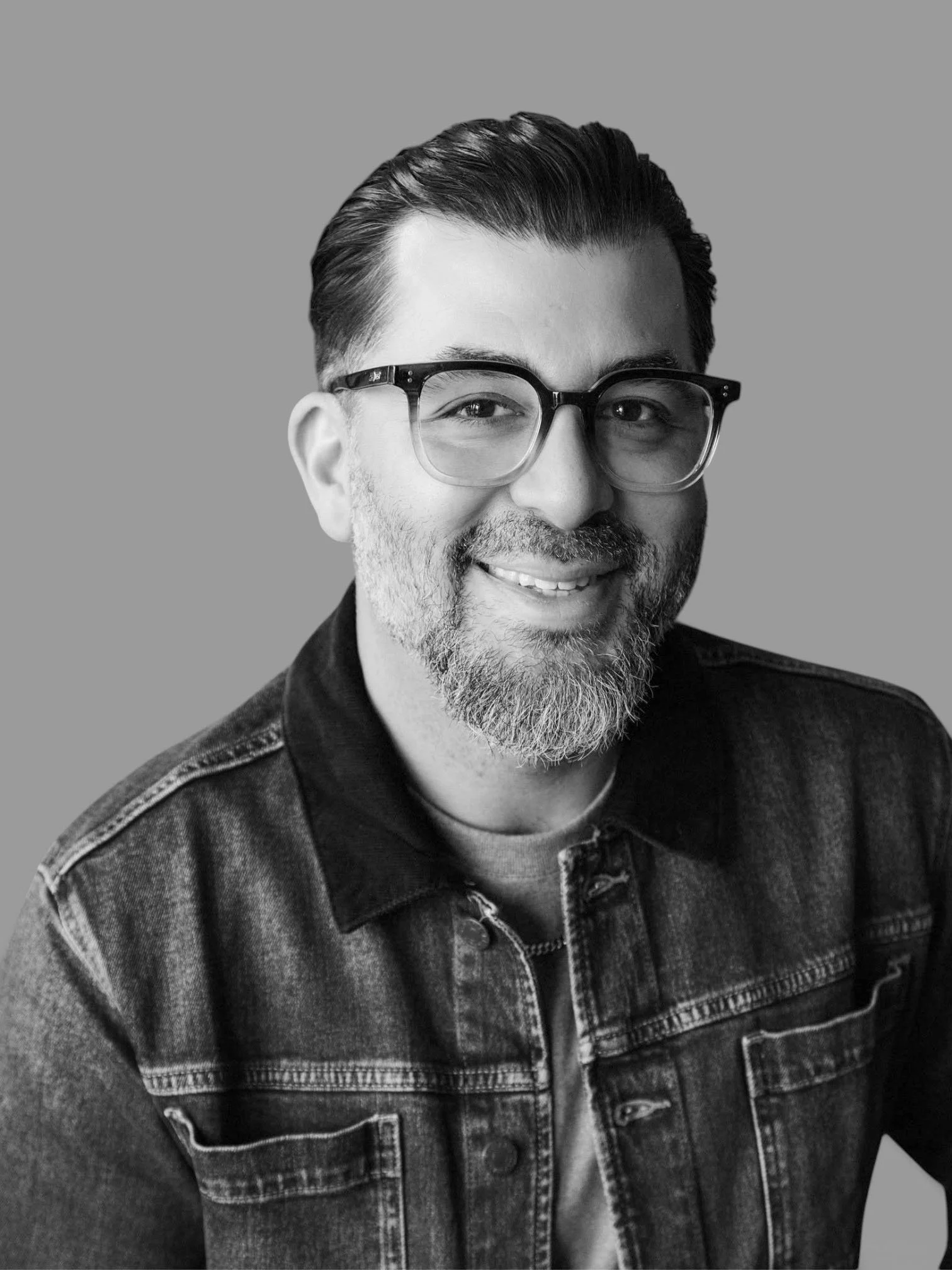 Black and white photo of a man smiling, wearing glasses and a denim jacket.