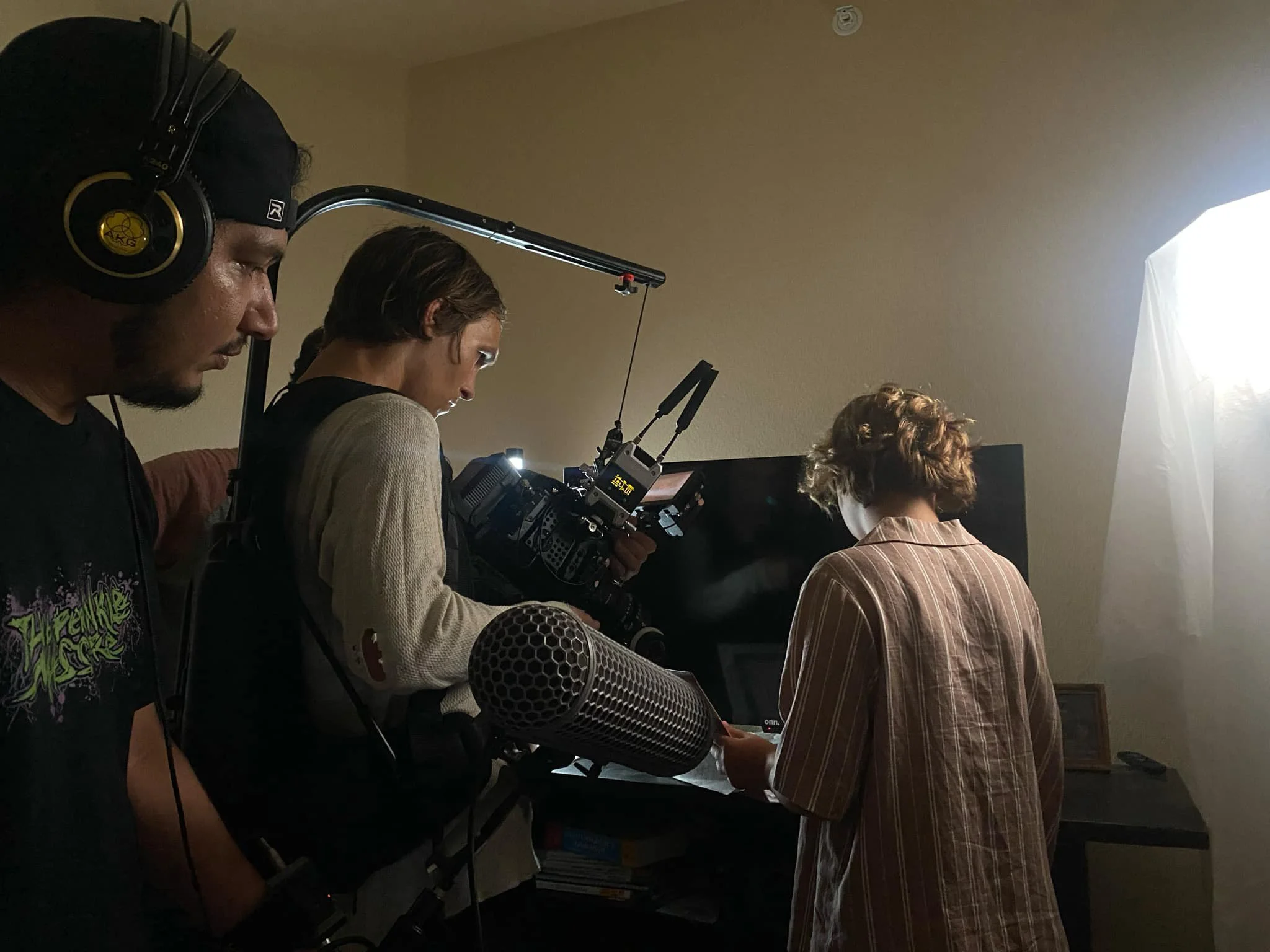 Corpus Christi Film crew members working with camera and equipment in indoor setting, with a person in a striped blazer and curly hair facing away from the camera, bright light source on the right side of the image.