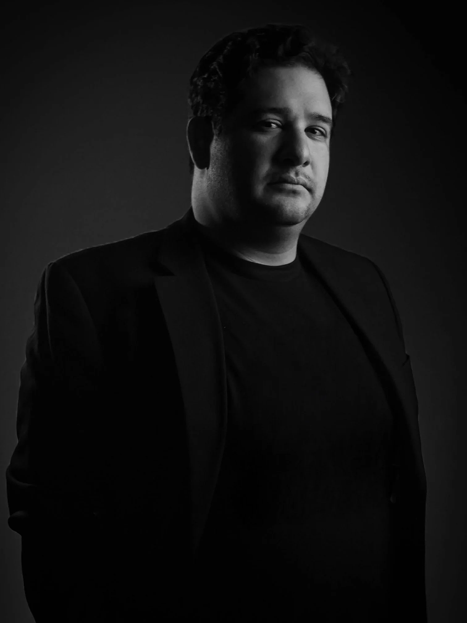 A black and white portrait of a man with dark, curly hair, wearing a black blazer and shirt, against a dark background.