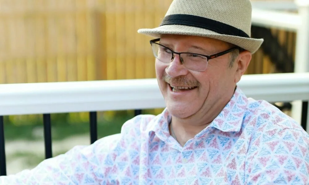 A man with glasses, a mustache, and a smile wearing a hat and a patterned shirt sitting outdoors near a wooden fence.