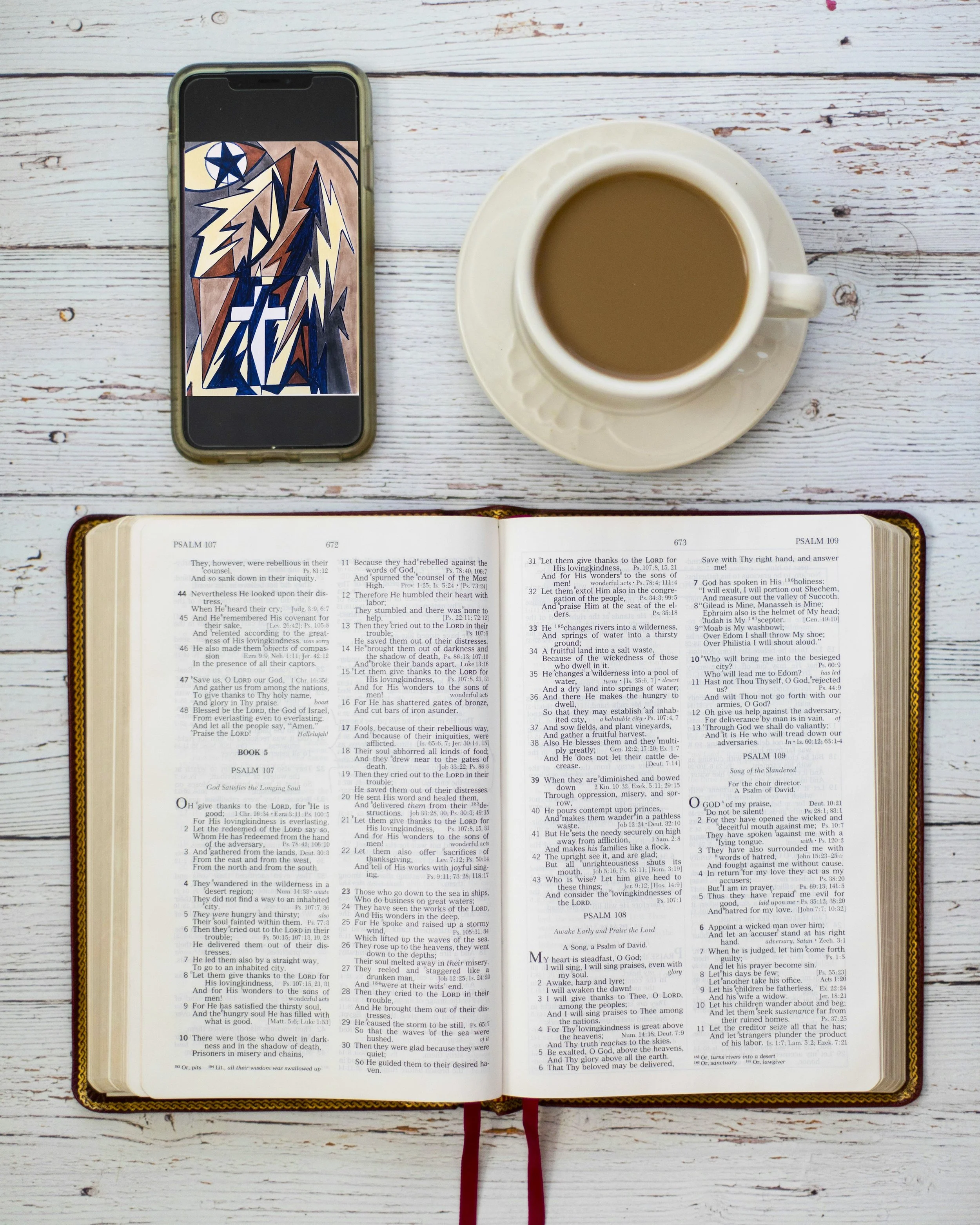 Open Bible with Psalm passages, a white cup filled with coffee on a saucer, a smartphone displaying abstract Cross Art artwork, all on a white wooden table.