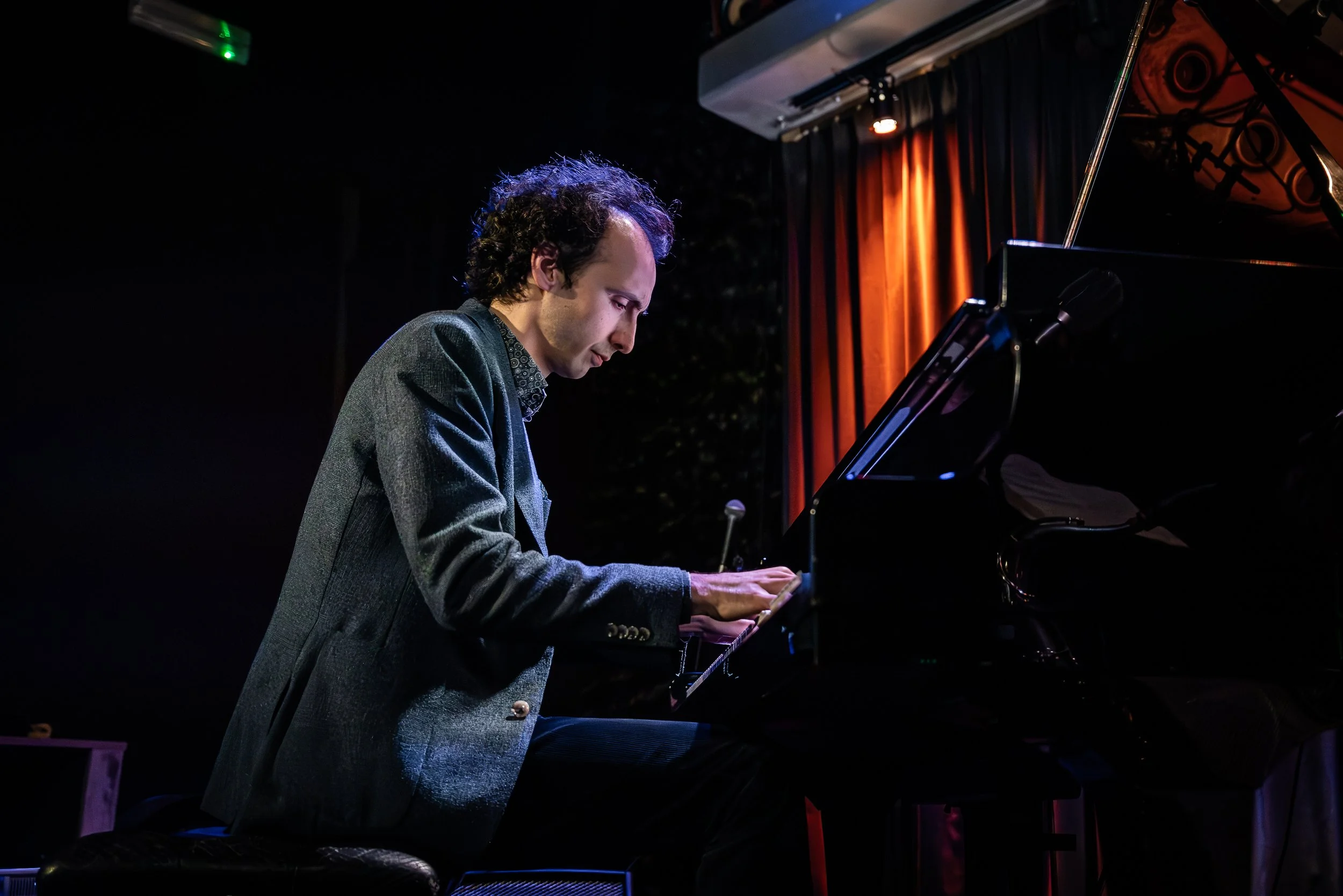 A man with curly hair wearing a gray blazer playing a grand piano on a dimly lit stage with orange curtains in the background.