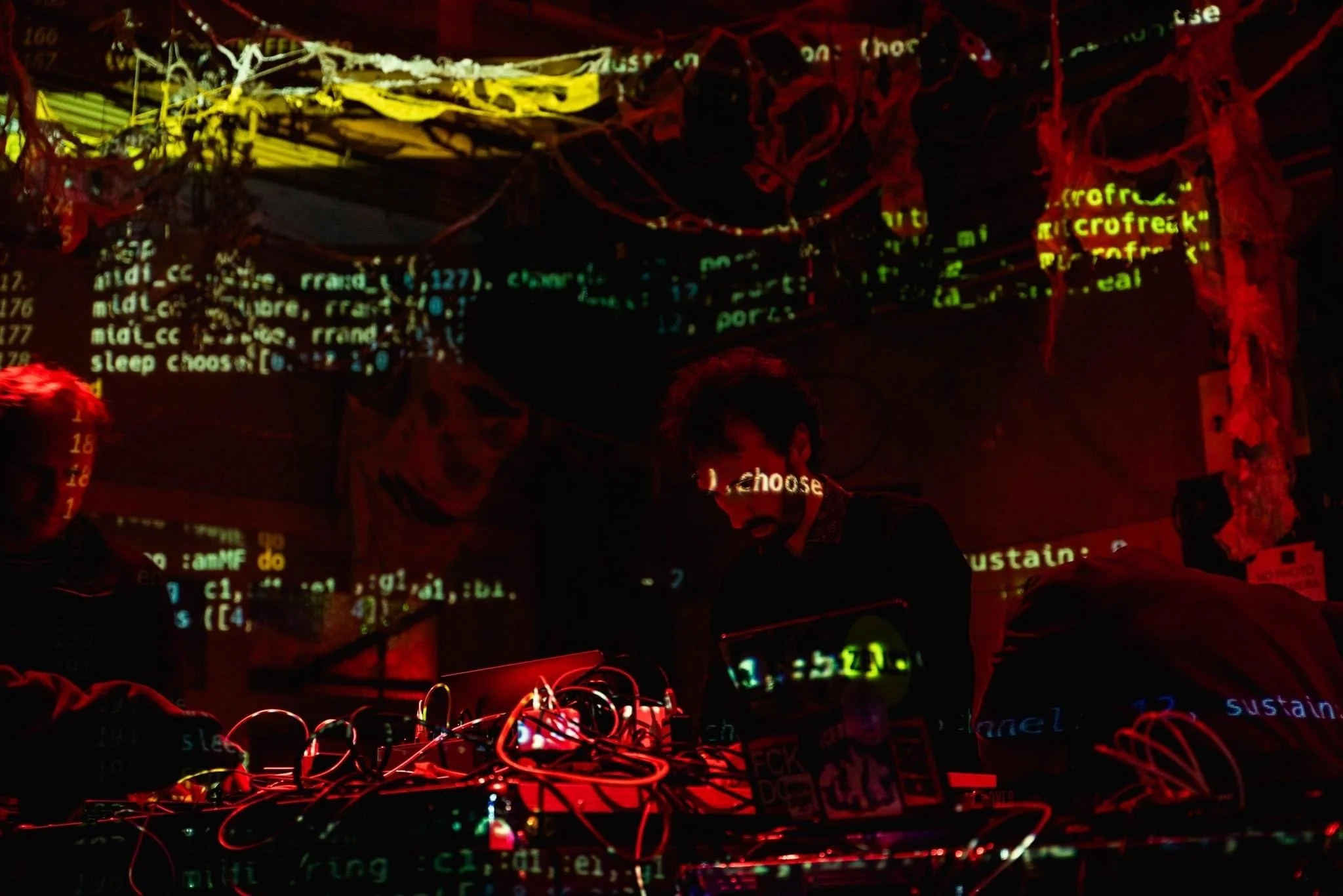 People in a dark room with colorful computer code and wiring projected on the walls, possibly a digital art installation or electronic music performance.