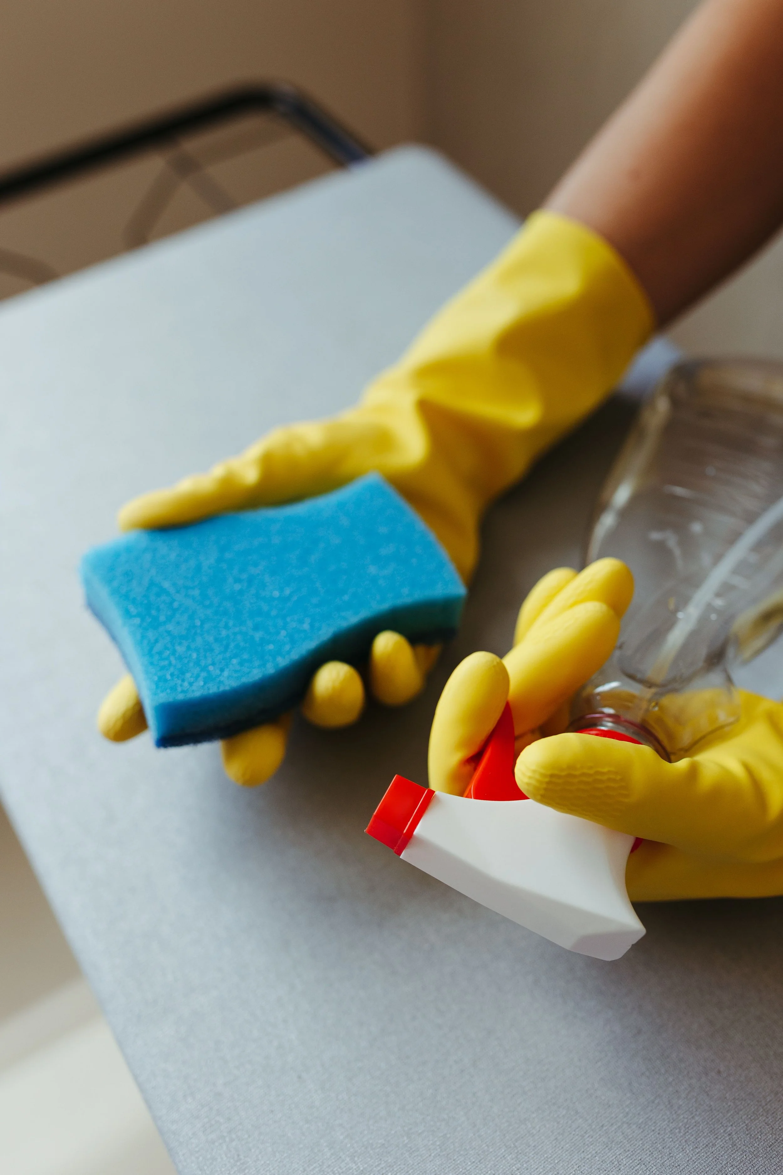A person wearing yellow rubber gloves holding a cleaning sponge and a spray bottle of cleaning solution, with cleaning supplies on a white surface.