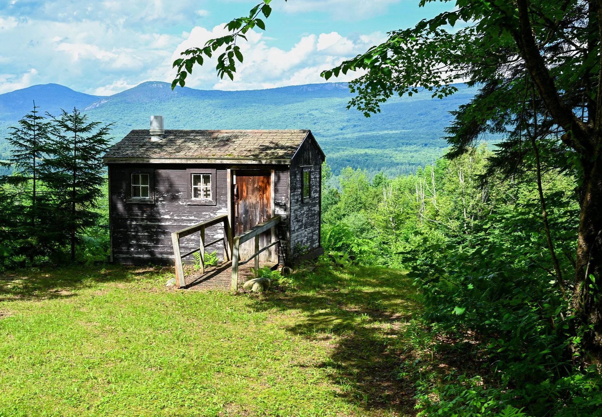A small, weathered black wooden house with a rusty metal door, set in a lush green forest with a mountain range in the background and partly cloudy sky.