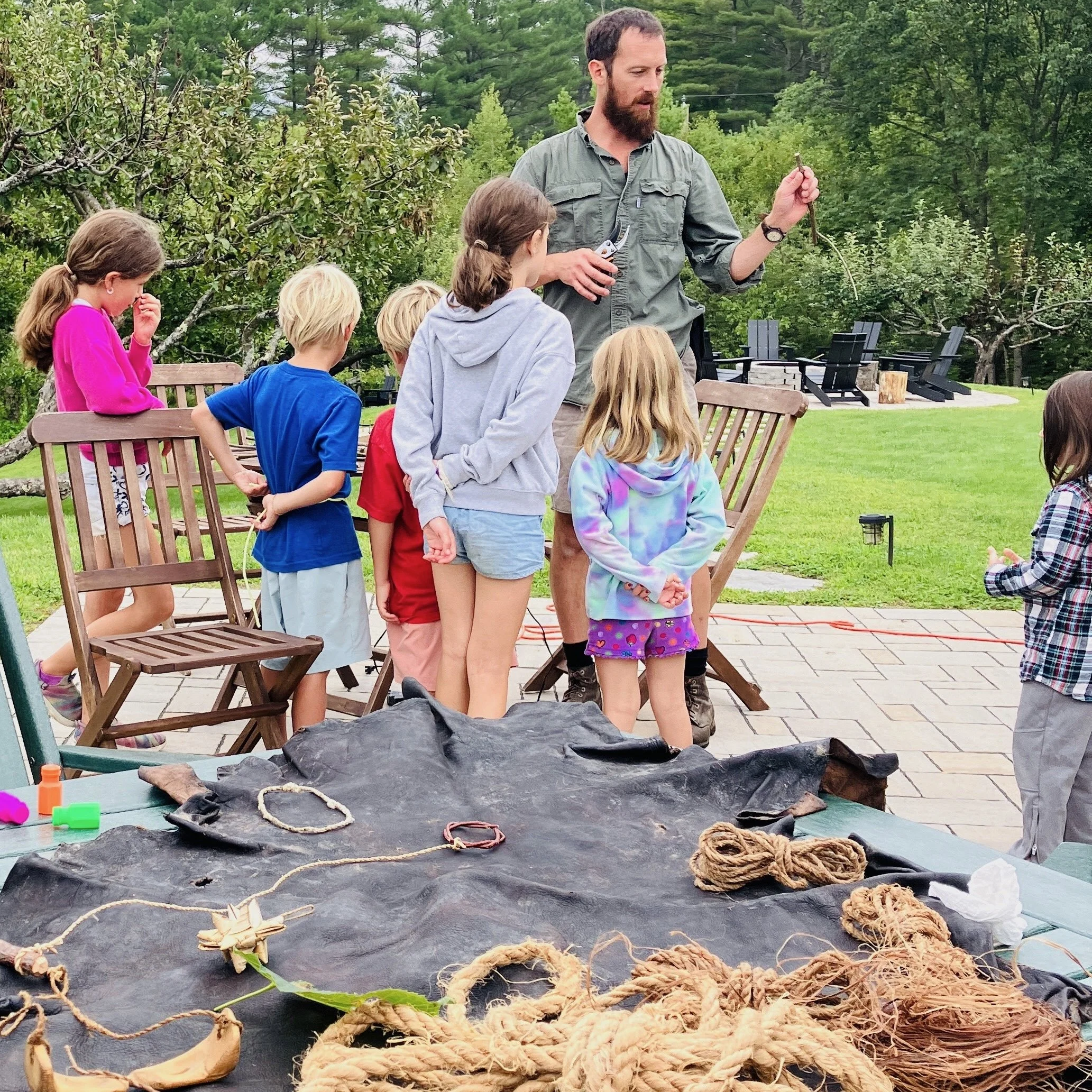 A man with a beard and gray shirt showing a rope to a group of children gathered outdoors on a patio or backyard, with trees and outdoor furniture in the background. The table in the foreground has ropes and tied knots.