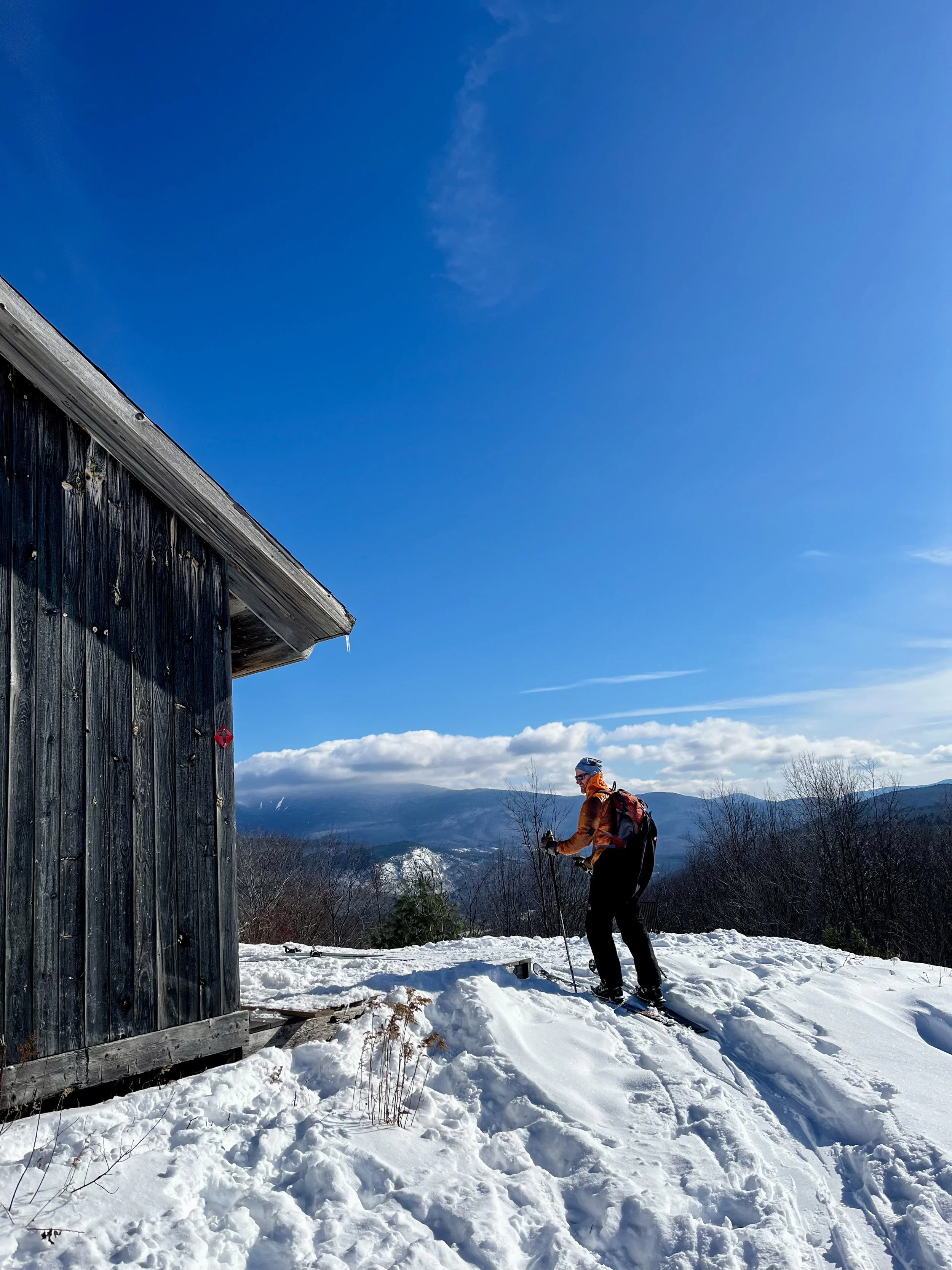 A person dressed for winter with skis and poles, standing outside near a wooden building with snow on the ground and a mountain view in the background.