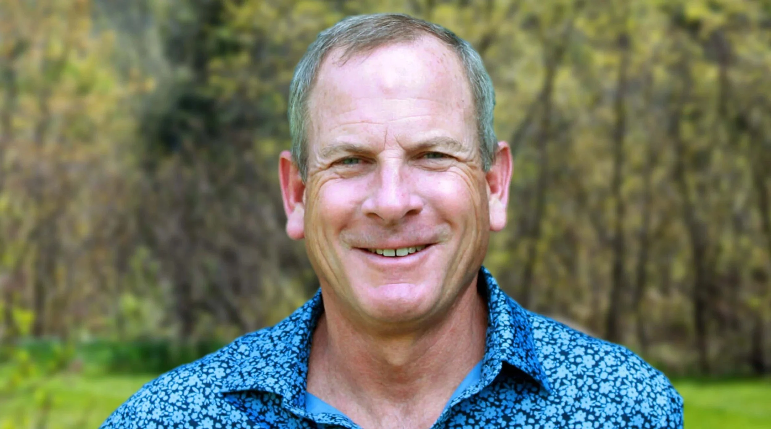 Portrait of a middle-aged man smiling outdoors, wearing a blue patterned shirt, with a background of trees and greenery.