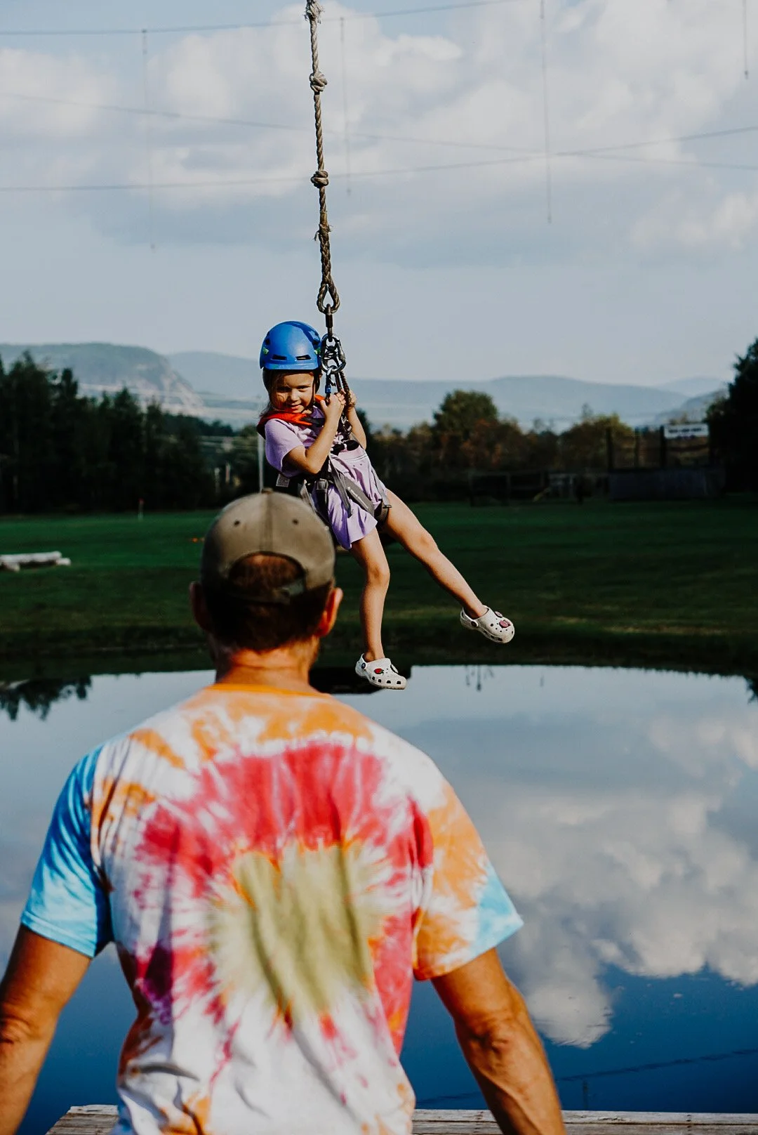 A young girl on a swing wearing a helmet, being watched by an adult. The scene is outdoors near a body of water, with hills and trees in the background.