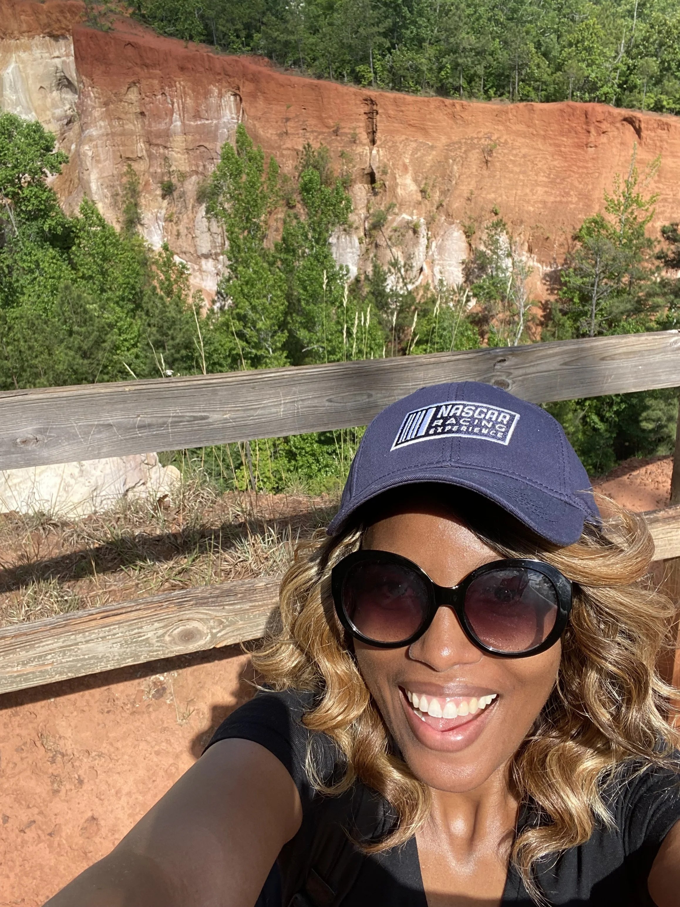 A woman taking a selfie outdoors, smiling, wearing sunglasses, a navy cap with NASCAR logo, and black shirt, standing in front of a wooden fence with a red cliff and green trees in the background at Providence Canyon State Park in Stewart County, GA.