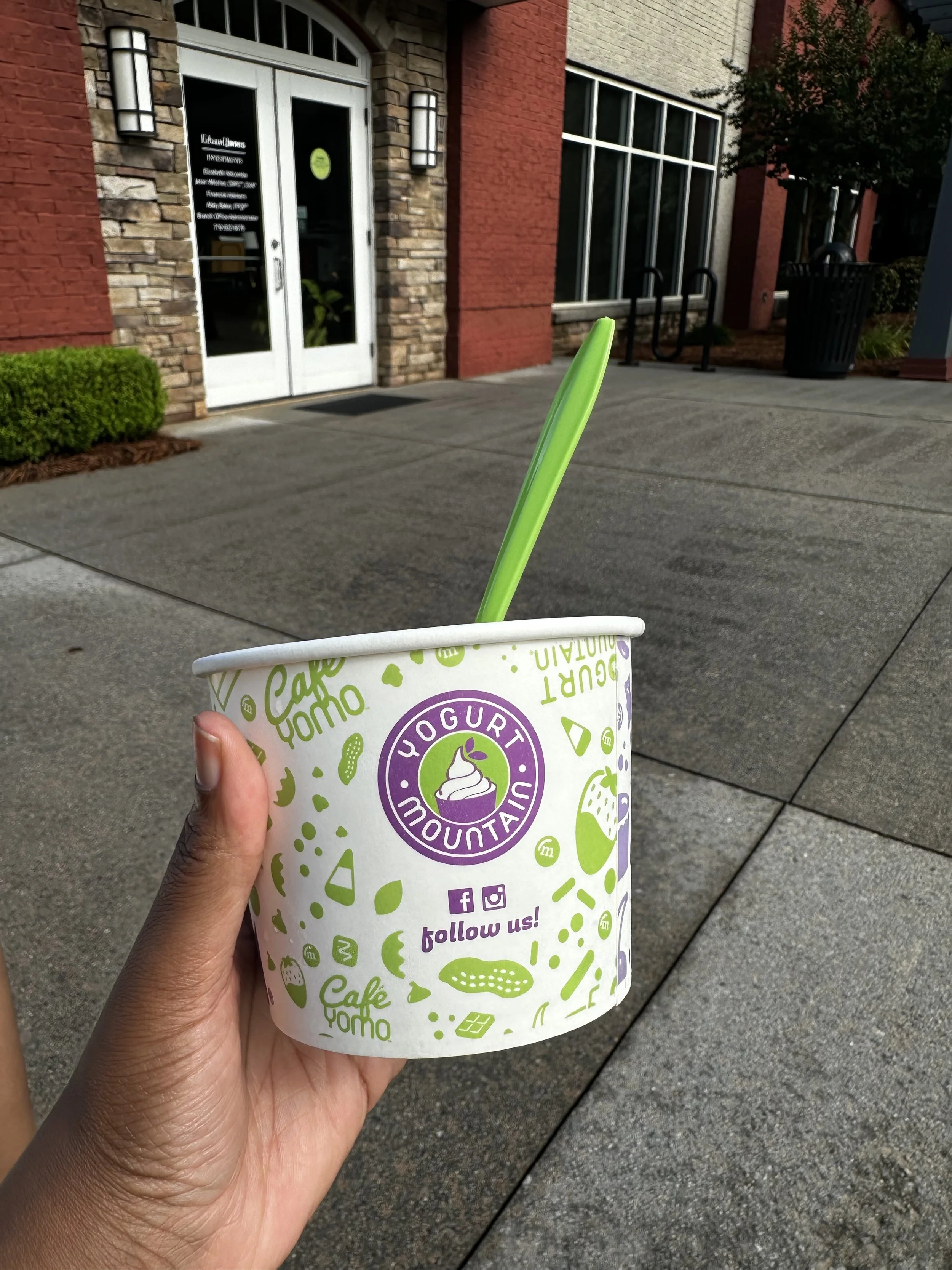 A person holding a cup of frozen yogurt from Yogurt Mountain in Conyers, GA with a green spoon outside a building with brick and stone walls, glass door, and large windows.