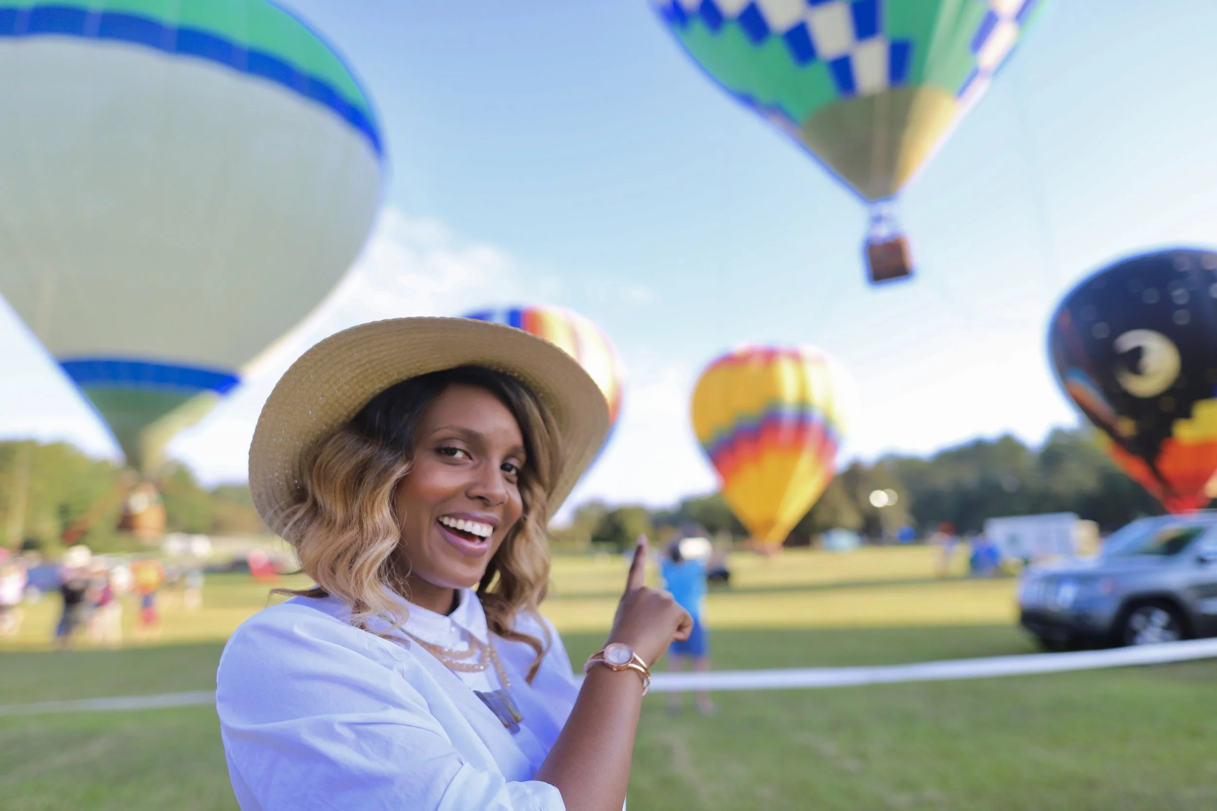 Sharmelle Brooks wearing a straw hat smiling and pointing at colorful hot air balloons in the sky during a festival in Pine Mountain, GA.