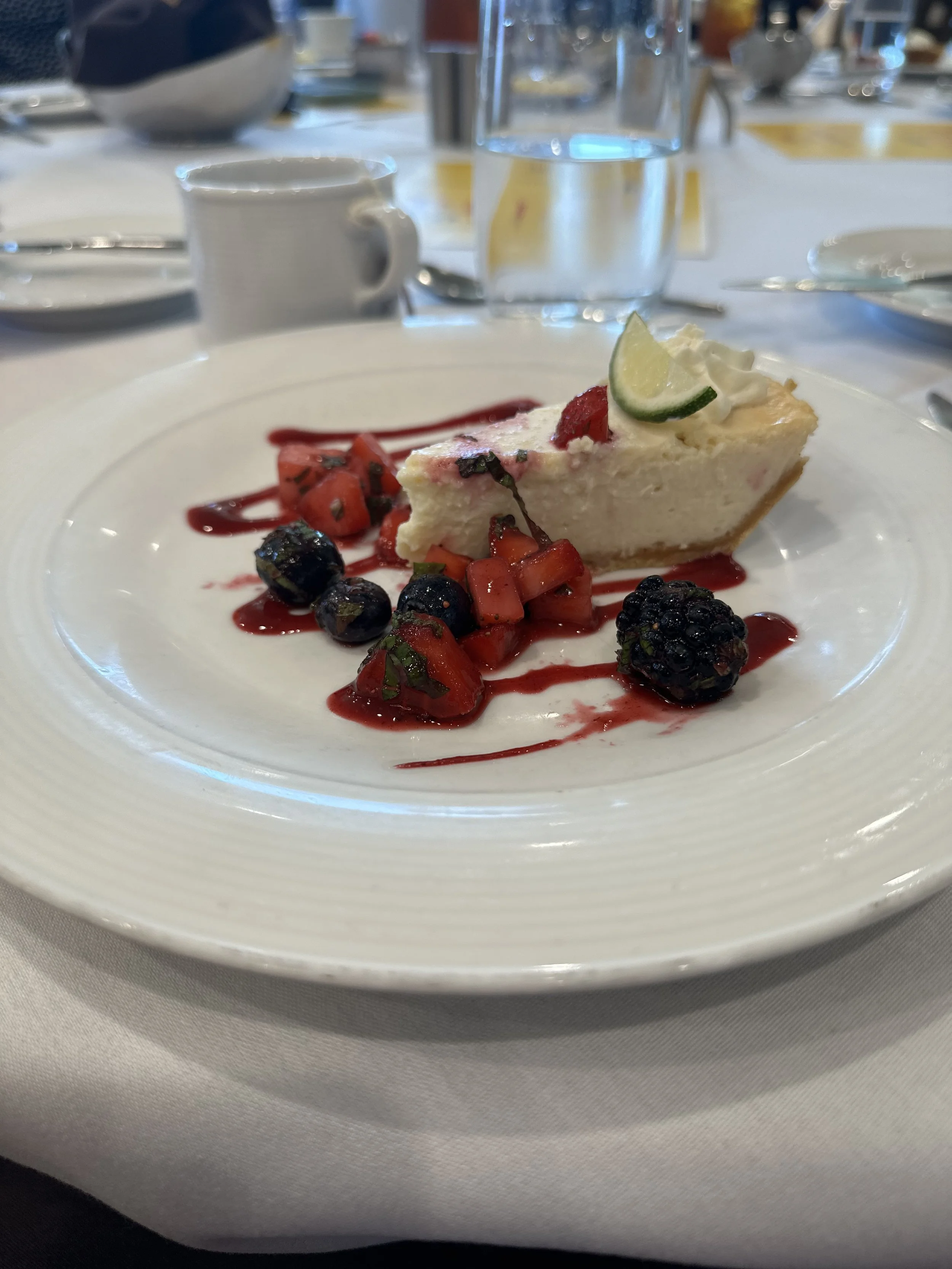 Slice of cheesecake with a lime wedge on top, garnished with mixed berries and a berry sauce drizzle, on a white plate.