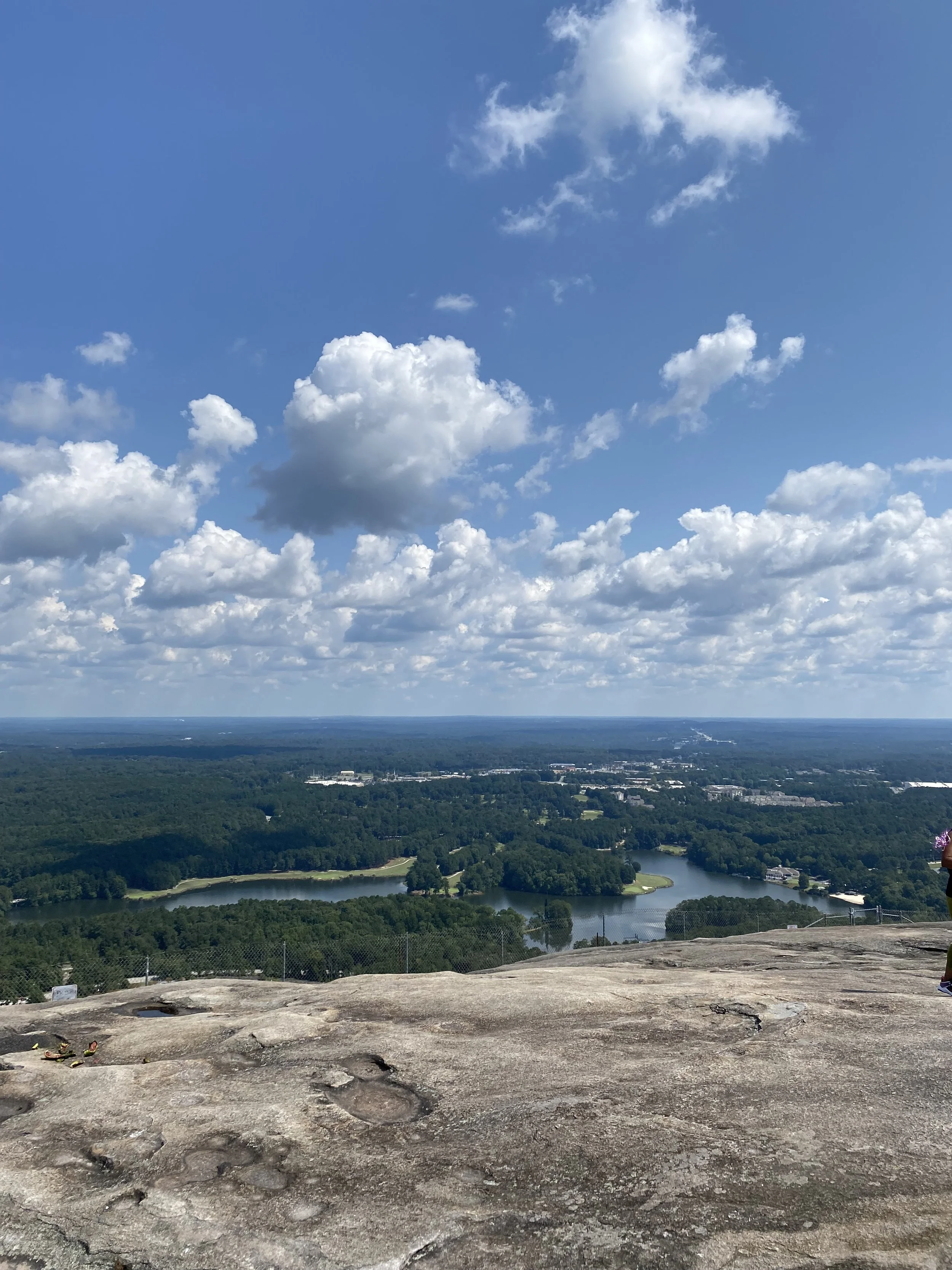 A scenic view of a vast landscape with a clear blue sky, white clouds, a winding river, lush green trees, and buildings in the distance, taken from a high vantage point with a rocky foreground at Stone Mountain Park, GA.