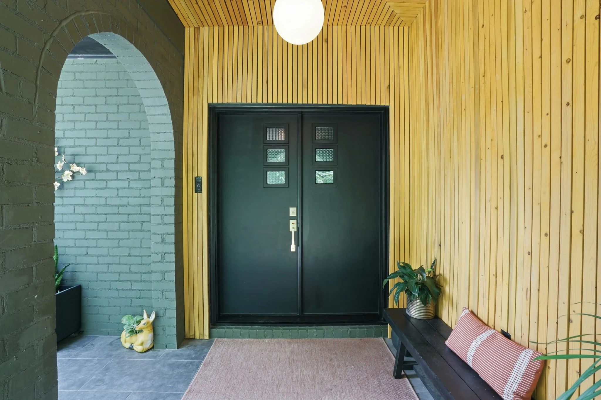 Entryway of mid-century modern home with black door, wooden wall paneling, houseplants, house shoes, and a bench with a pillow in Smyrna, GA.