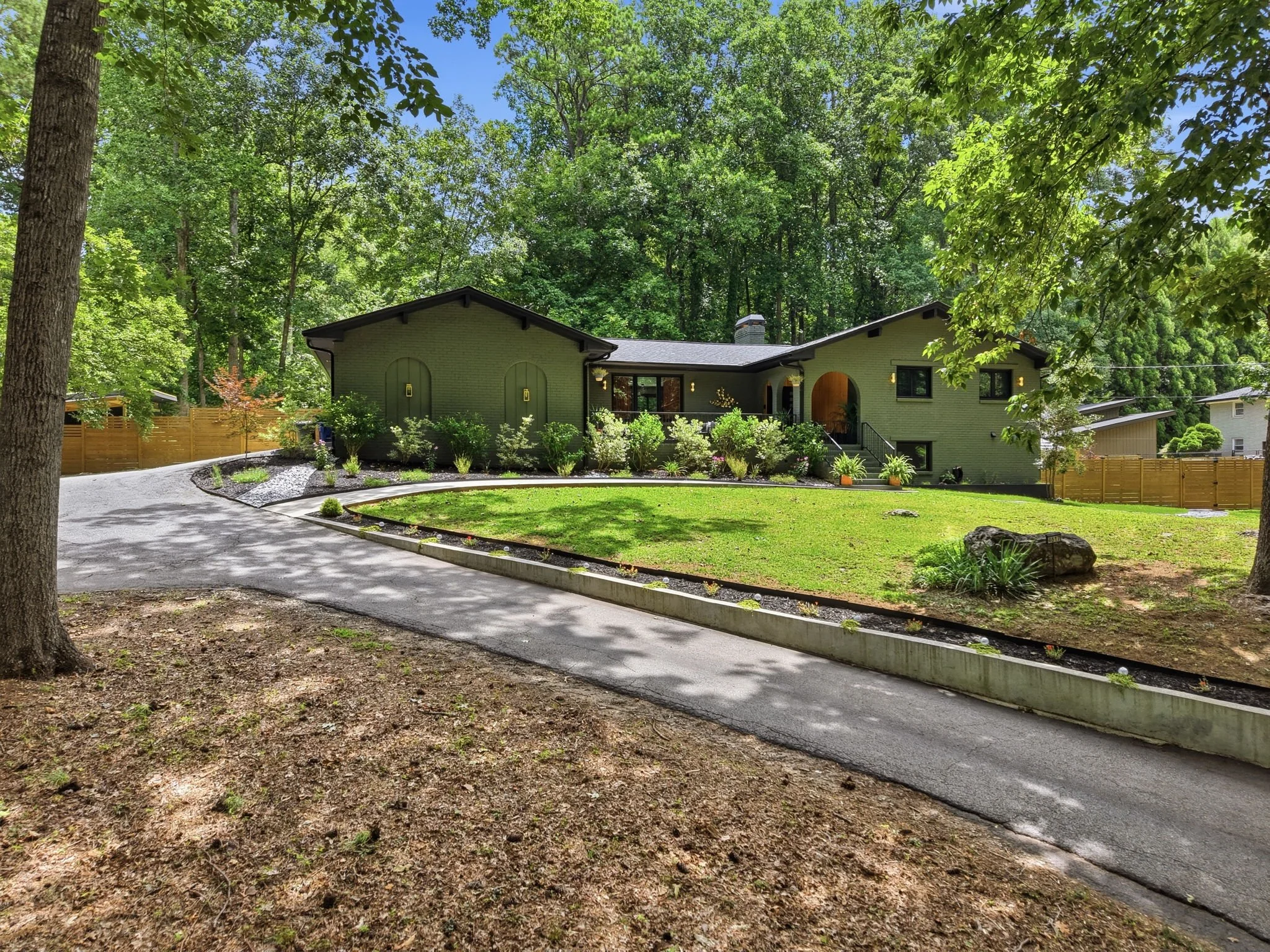 A renovated mid-century modern home surrounded by tall green trees with a front lawn, a curved driveway, and plants along the house. The house is painted dark green with black roof accents in Smyrna, GA.
