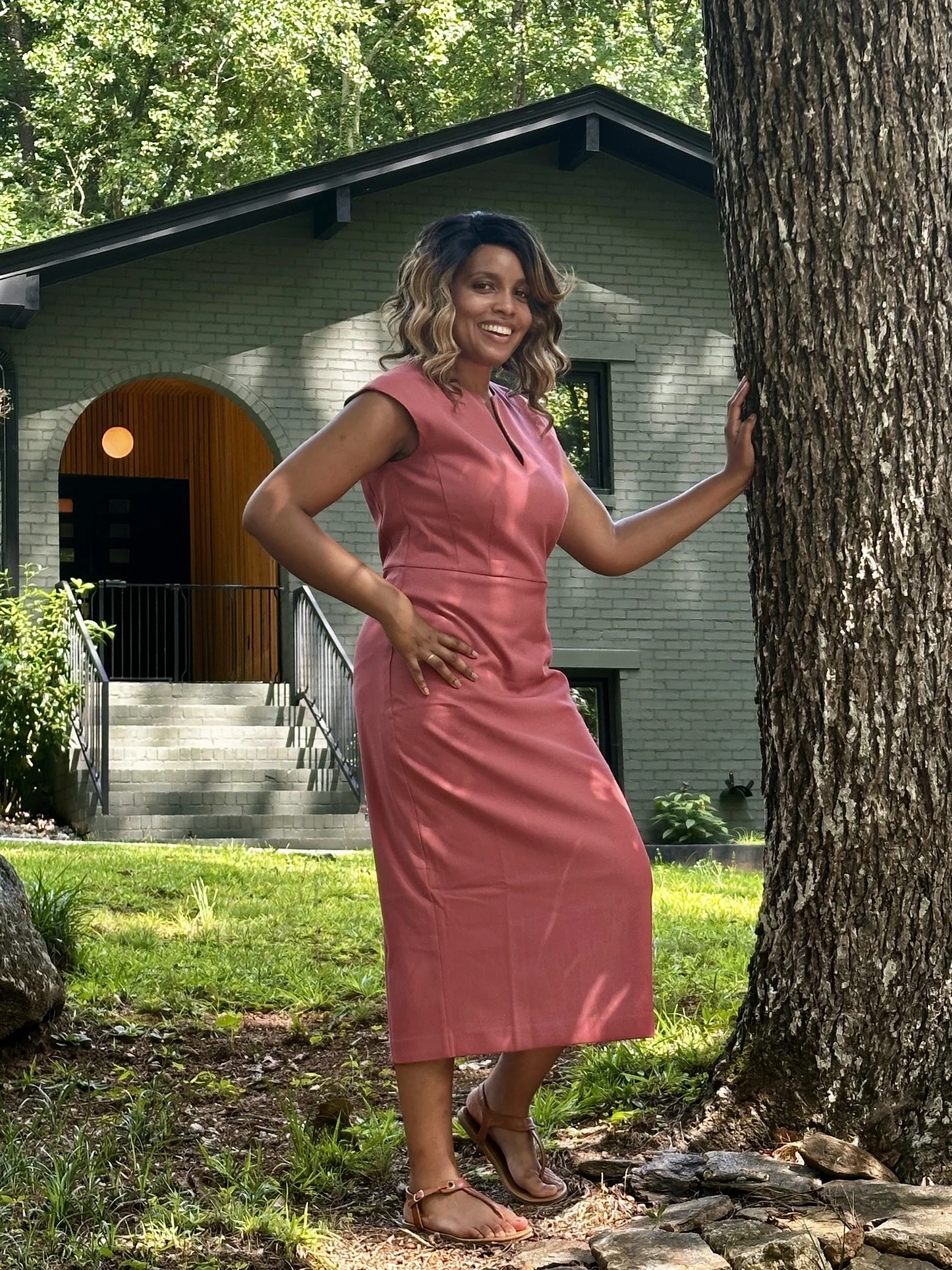 Sharmelle Brooks, metro Atlanta REALTOR and Associate Broker wearing a pink dress standing outdoors near trees and a house, leaning against a tree with one hand on her hip and the other on the tree trunk, smiling at the camera in Smyrna, GA.