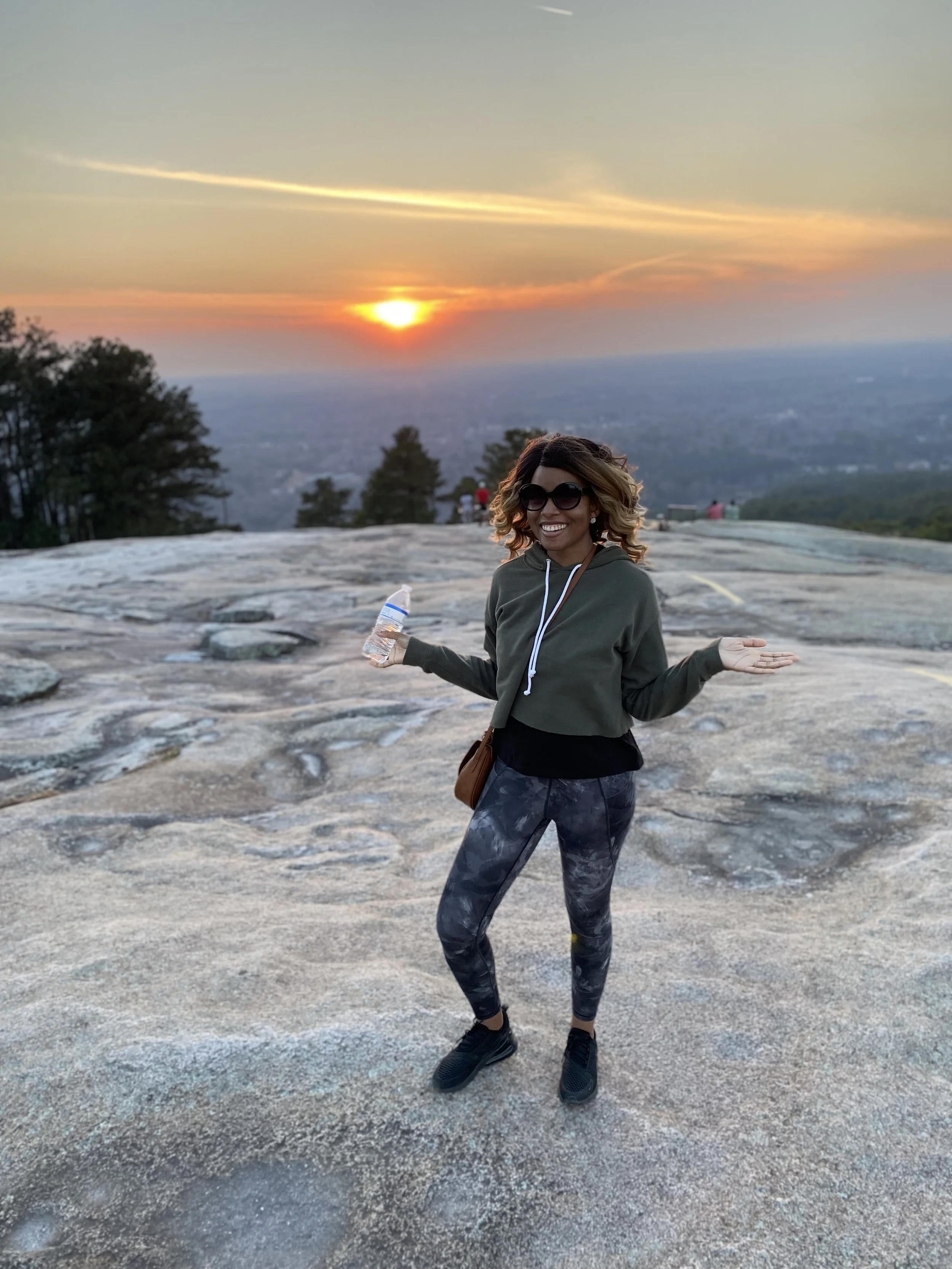 A woman with curly blond hair, wearing sunglasses, a green hoodie, and patterned leggings, smiling with arms outstretched while standing on a rocky area during sunset, with trees and a city view in the background at Stone Mountain Park, GA.