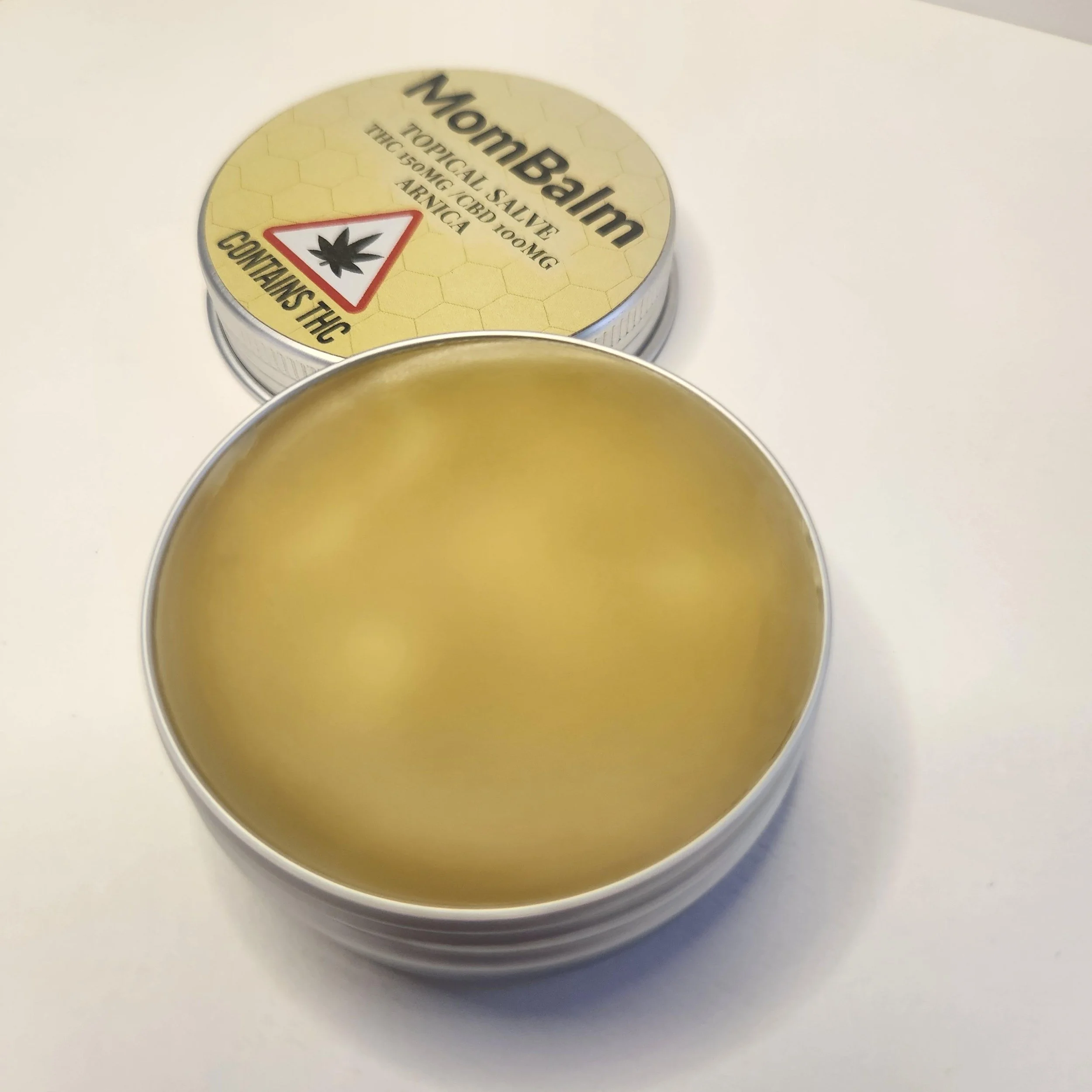 Close-up of a glass jar lid with a label that says 'Hemp Balm' and a warning symbol with a cannabis leaf inside a triangle. The label describes it as a topical salve with THC, 150 mg/CBD, 100 mg. The jar is placed on a white surface.