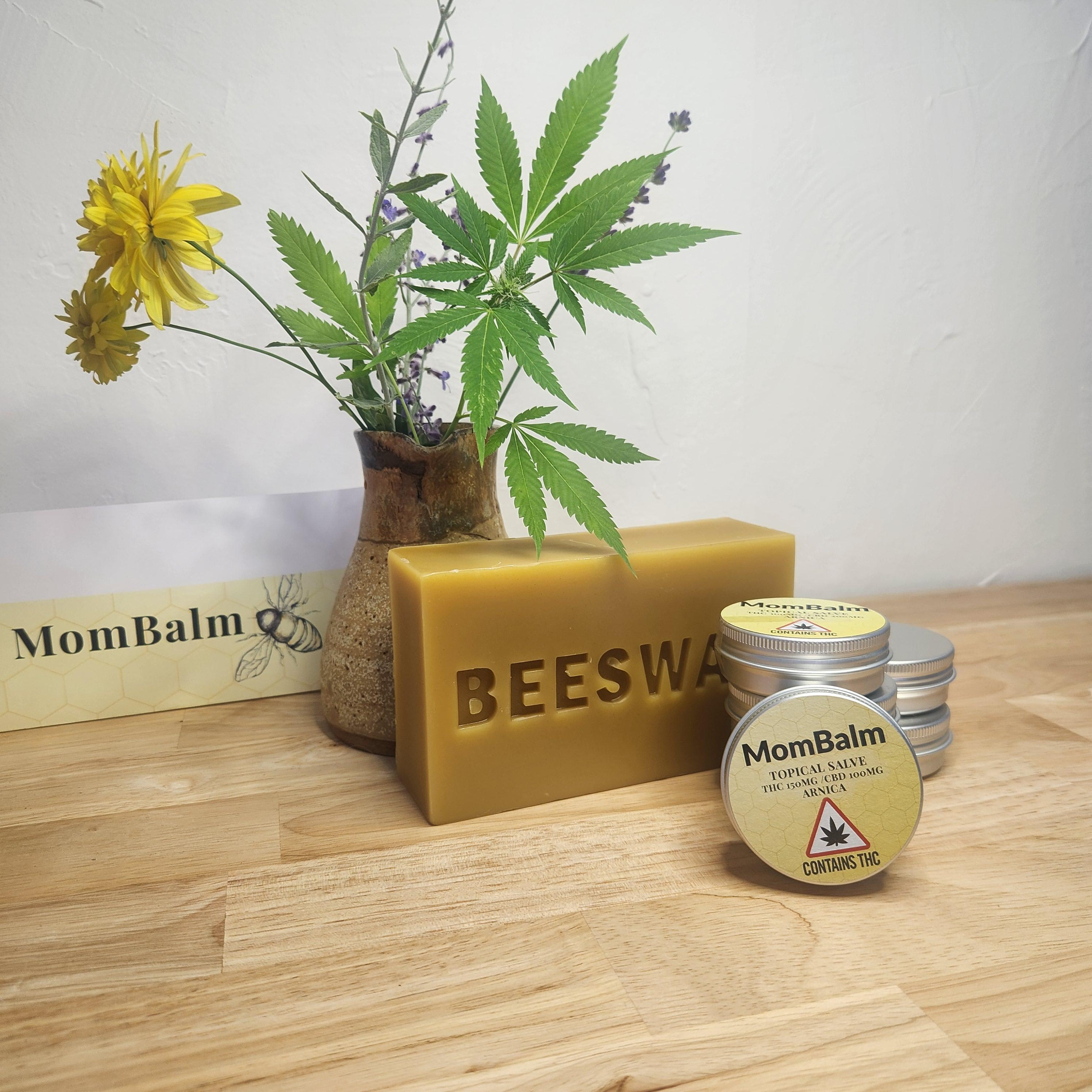 A plant with marijuana-like leaves in a pot, yellow flowers and purple small flowers, arranged among skincare and cannabis products on a wooden surface.