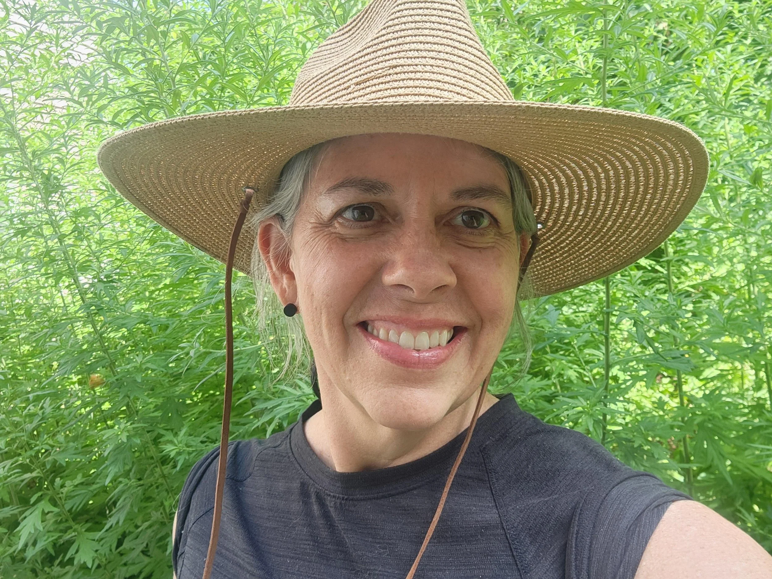 A smiling woman wearing a wide-brimmed straw hat and a dark gray shirt, standing outdoors among lush green foliage.
