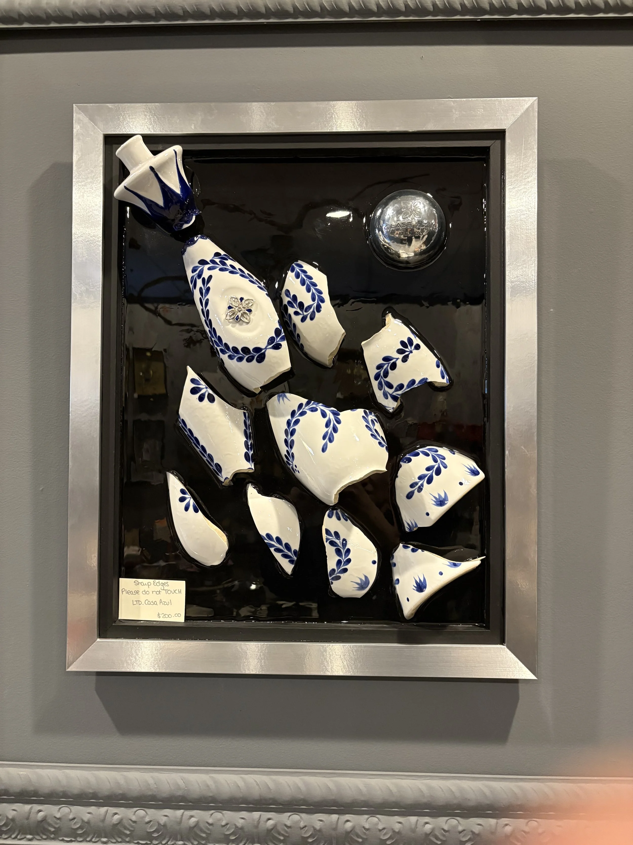 Decorative ceramic pieces with blue floral patterns arranged inside a black shadow box with a silver frame. Small note on the bottom left reads: 'Scrap edges Please do not TOUCH LTD. Casa Azul.'