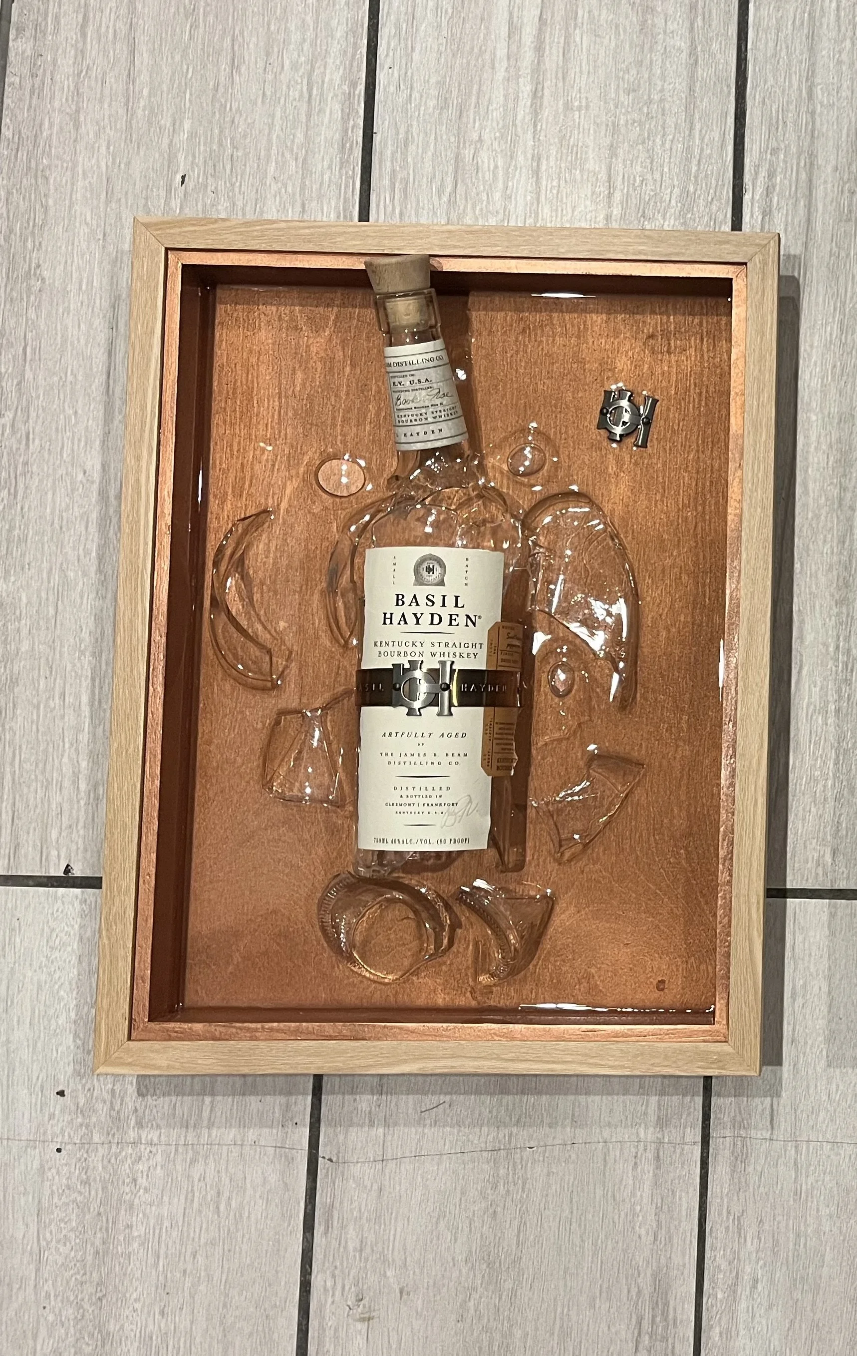 A glass bottle of Basil Hayden Kentucky Straight Bourbon Whiskey inside a shadow box frame with a wooden border. The bottle is surrounded by empty glass whiskey glasses and a metal clip.