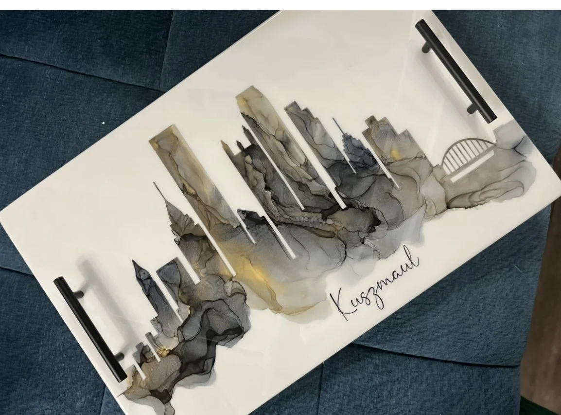 A decorative wall art piece with a cityscape silhouette featuring buildings, a bridge, and clouds in black, gray, and gold tones, with the word "Kusamallu" written below.