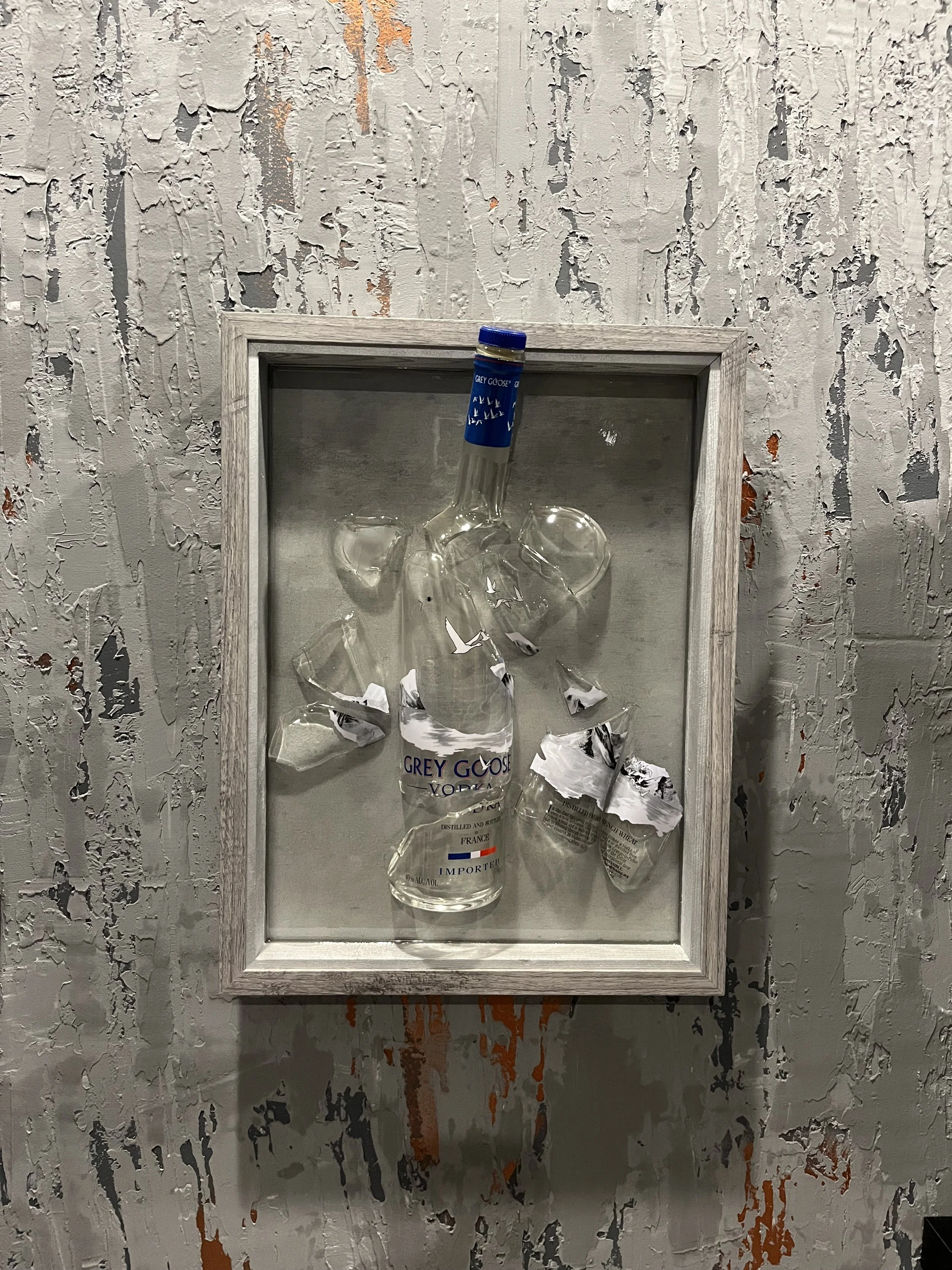 A framed display with broken glass and a bottle of Grey Goose vodka attached inside, mounted on a distressed, textured wall with peeling paint.