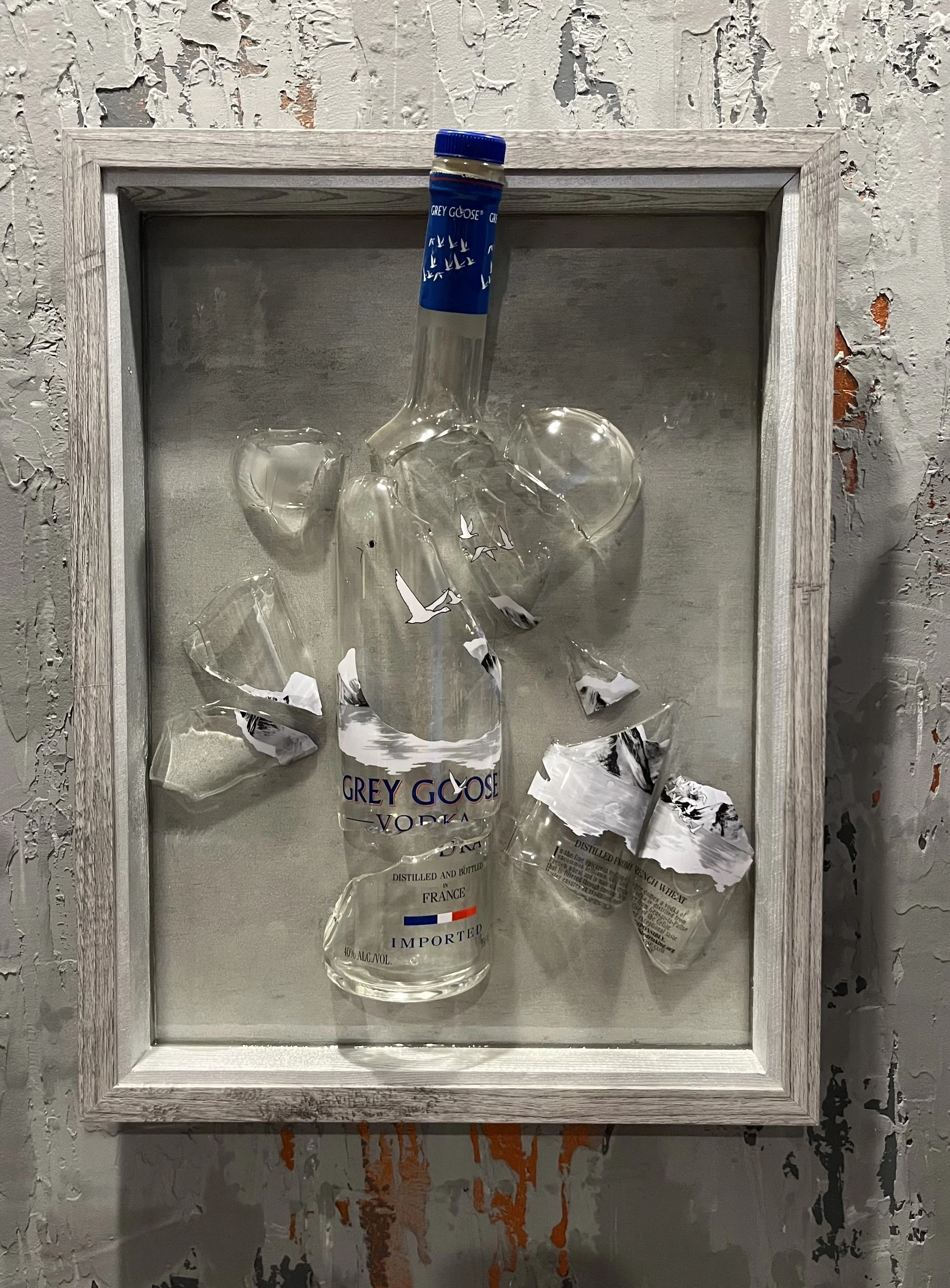 Framed display with broken glass containing a nearly empty Grey Goose vodka bottle and torn pieces of paper, set against a textured wall with chipped paint.
