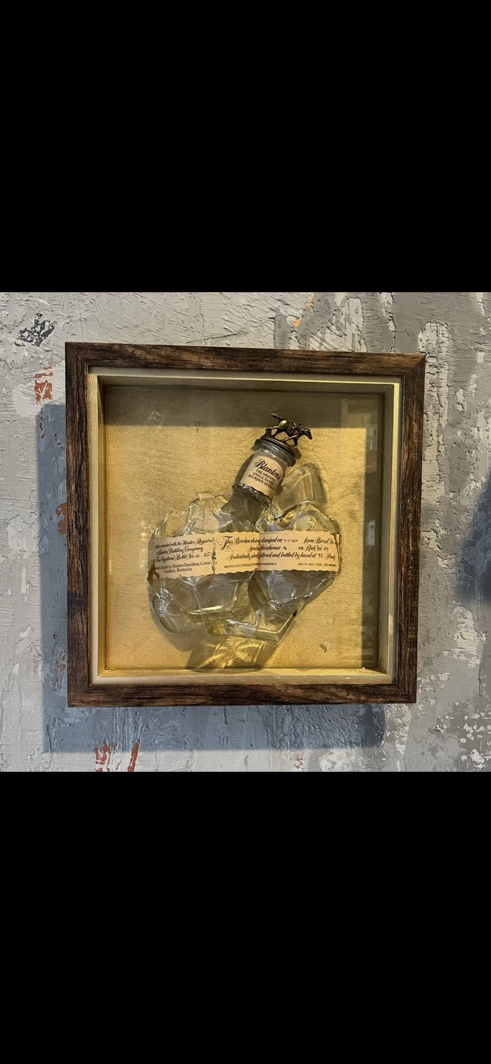 A framed shadow box display featuring a glass bottle with a small horse figurine on top, surrounded by crushed ice and a handwritten document or label inside the frame, mounted on a textured wall.