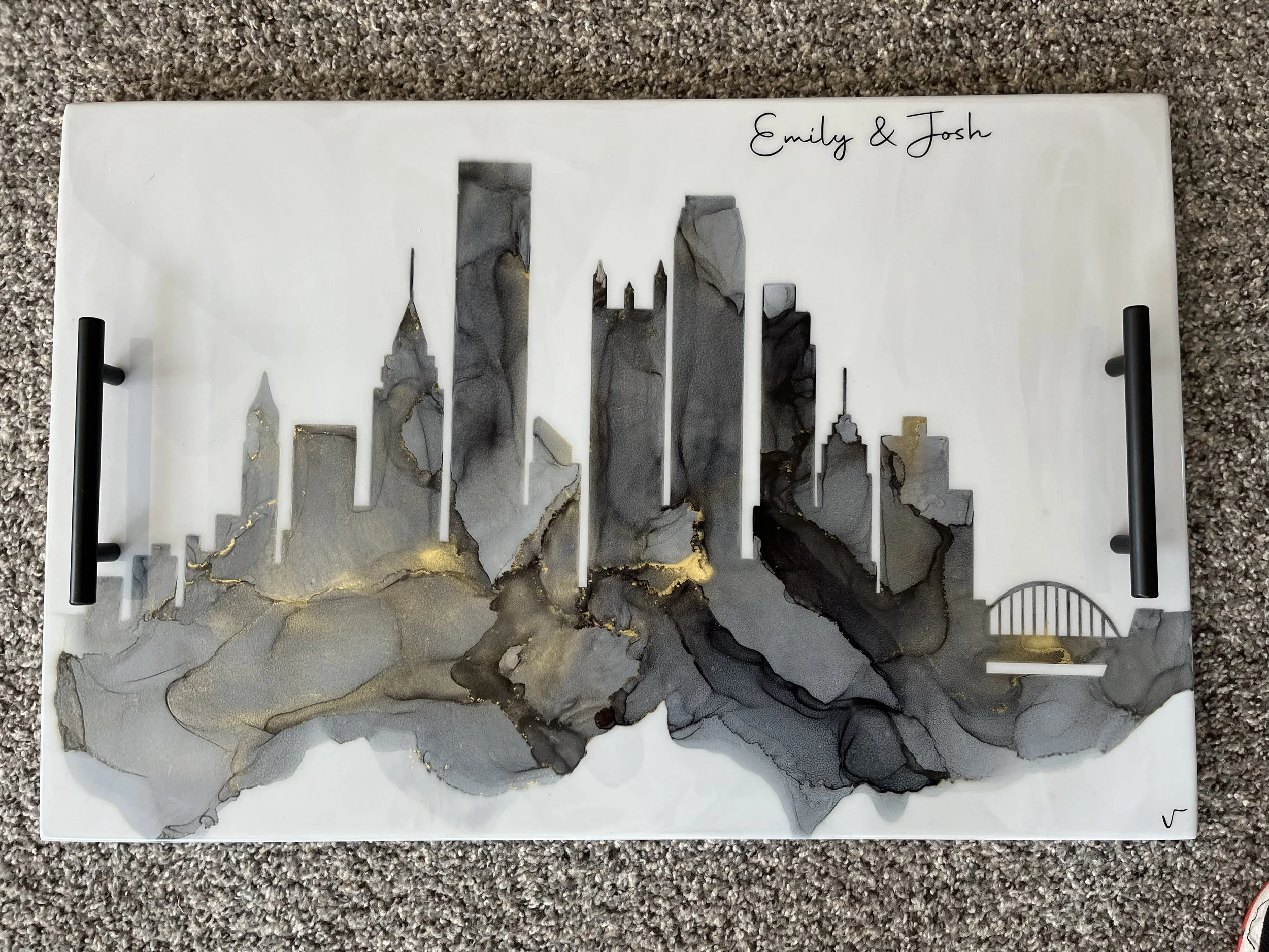A decorative wall art piece with a silhouette of a city skyline crafted from gray, black, and gold marbled resin, mounted on a white background with the names Emily and Josh written in black cursive at the top right corner.