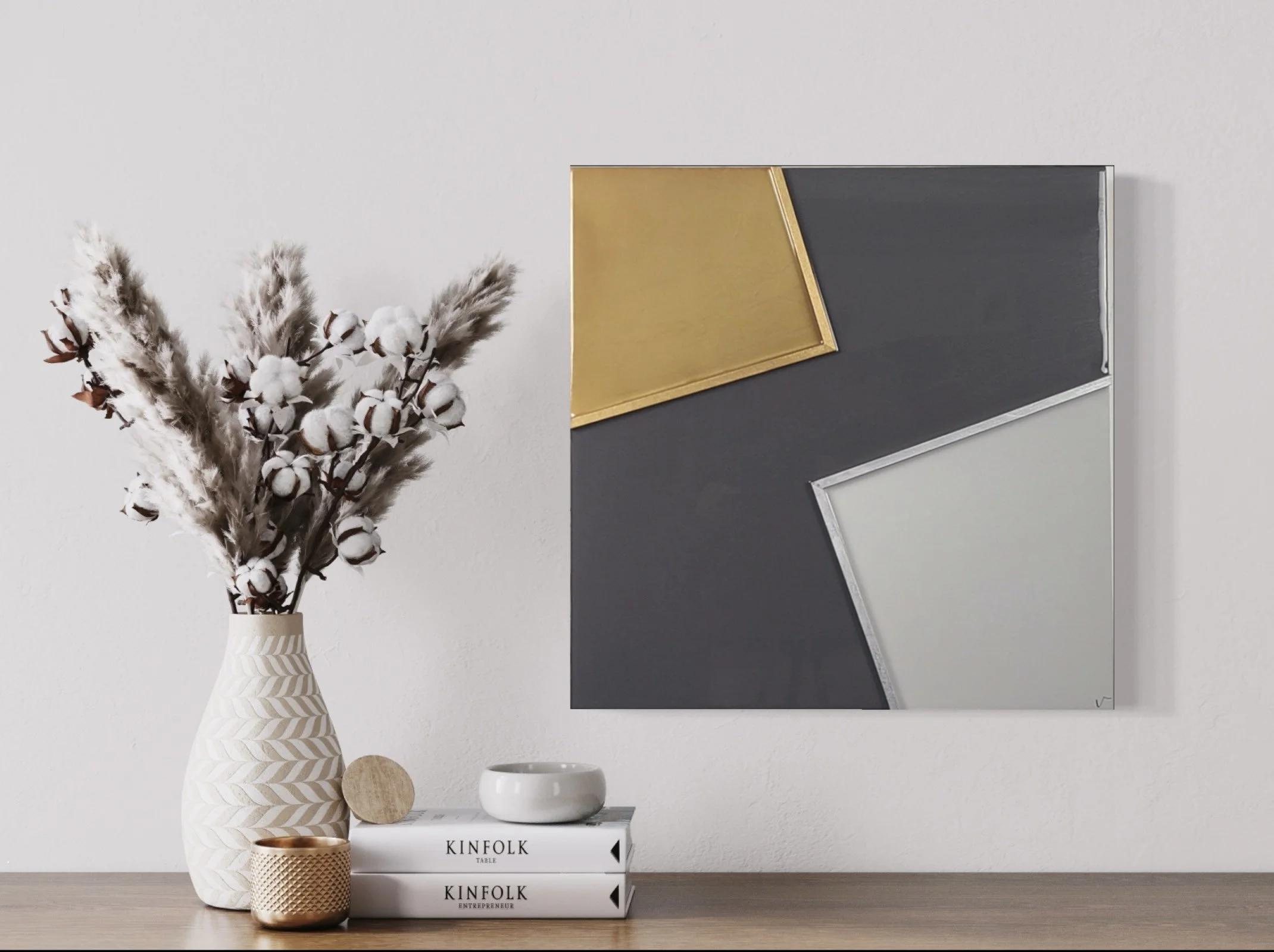 A decorative arrangement with dried flowers in a white vase with a gold chevron pattern, a smaller brown textured vase, and a white candle holder on top of stacked books titled 'KINFOLK TABLE', 'KINFOLK ENTREPRENEUR', with a modern abstract painting on a wall in the background.