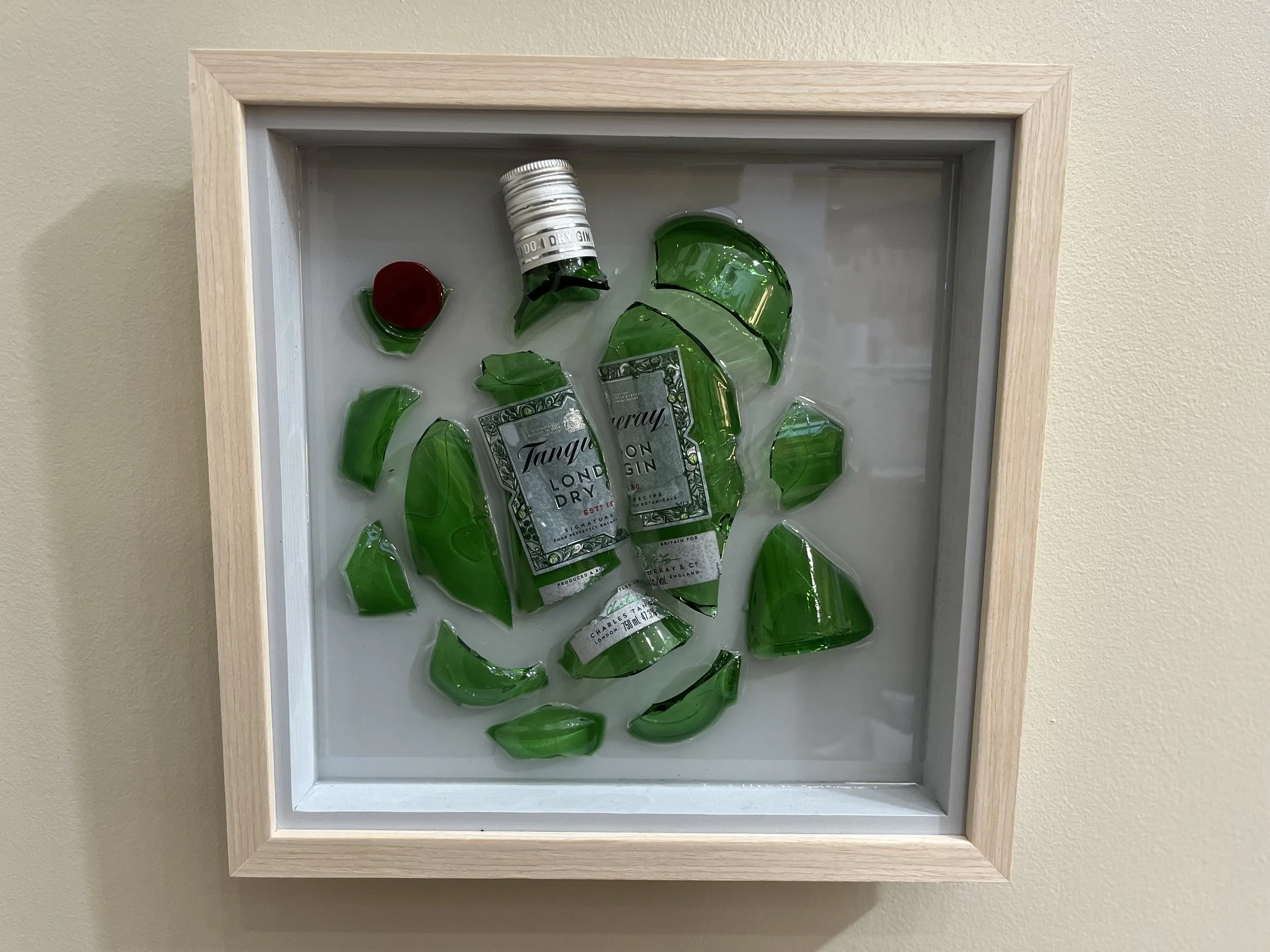 A shadow box containing broken green glass pieces, a small container with a red lid, and a label reading "Tanqueray".
