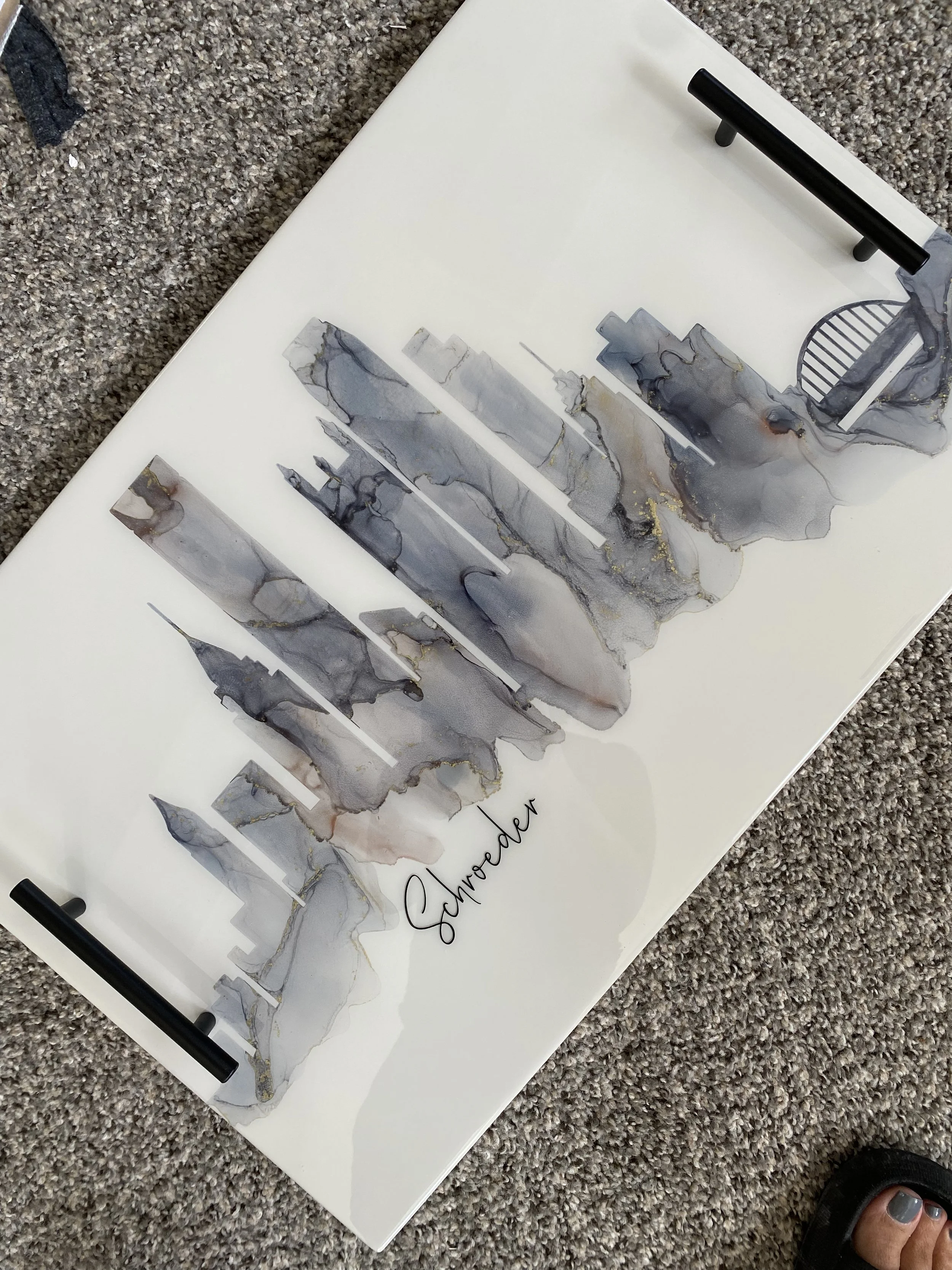 A white sign with a watercolor cityscape illustration in shades of gray, black, and gold, with the word "Savoreah" handwritten in black at the bottom. The sign is on a textured gray carpet, with black corner protectors and a person’s foot in a sandal