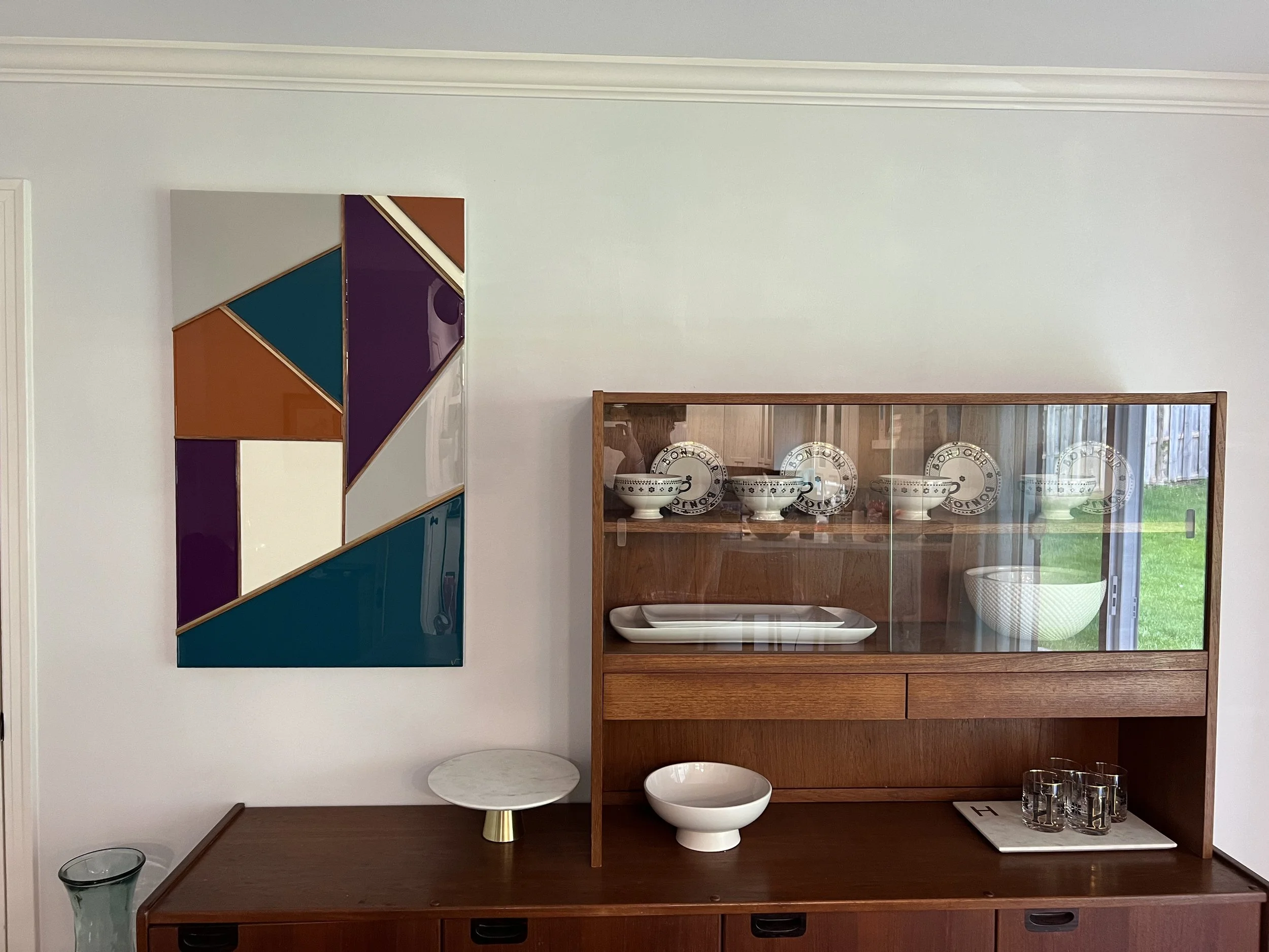A wooden display cabinet with glass doors showcasing bowls and plates, next to a wall with colorful geometric art, and a small white table and bowls on a dark wooden surface.